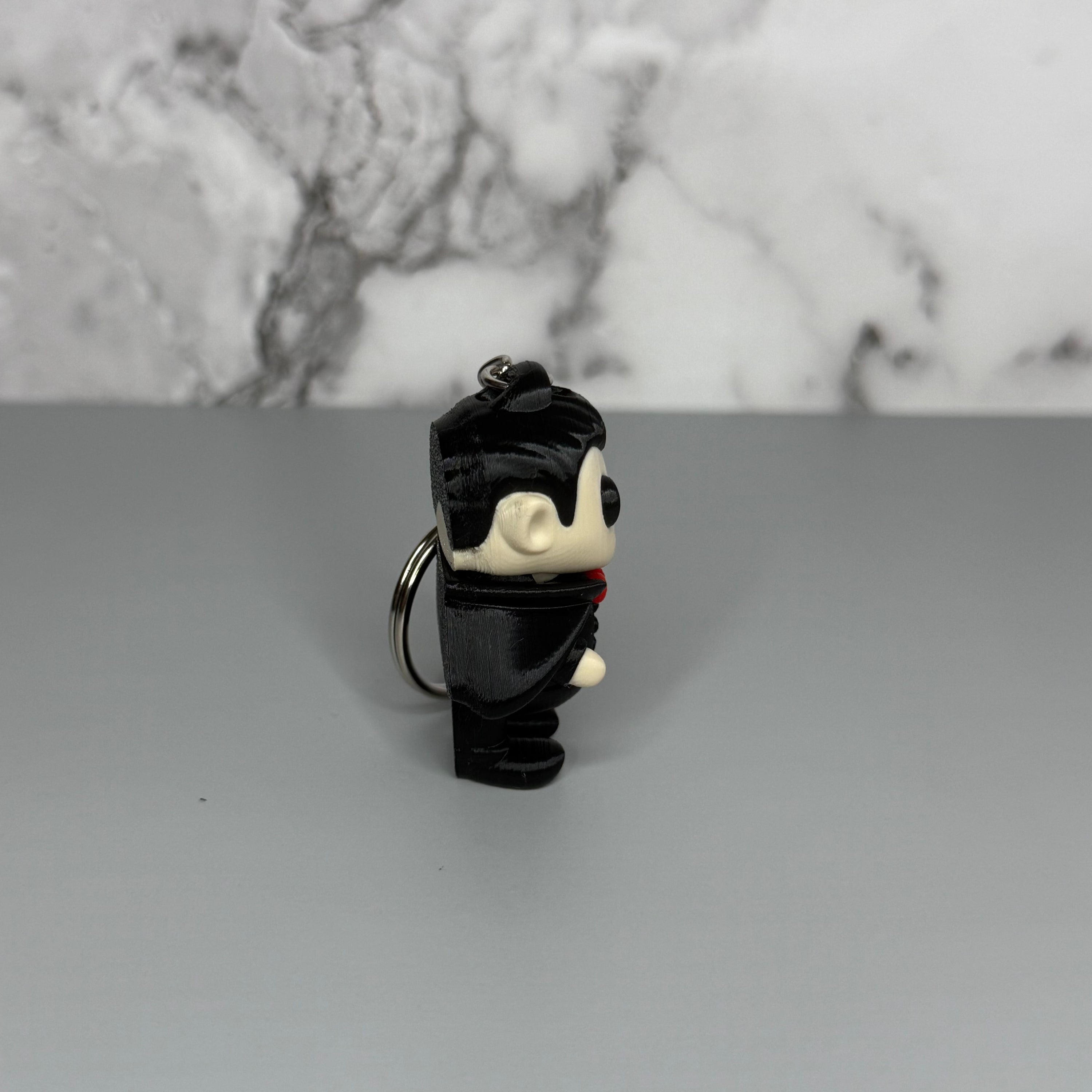 Left side of a keychain of a dracula vampire figure with a black cape, white fangs, and a red bowtie on a gray marble background