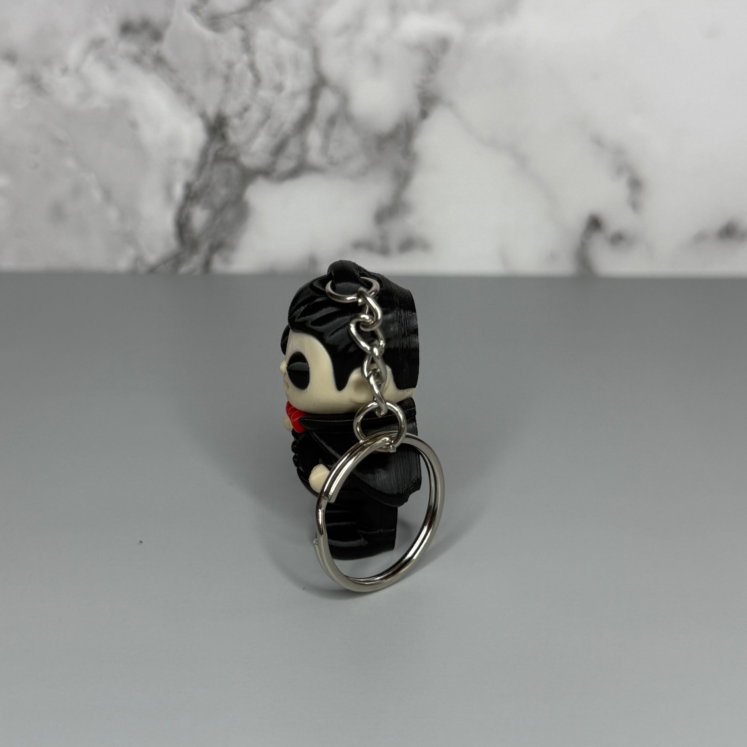 Right side of a keychain of a dracula vampire figure with a black cape, white fangs, and a red bowtie on a gray marble background
