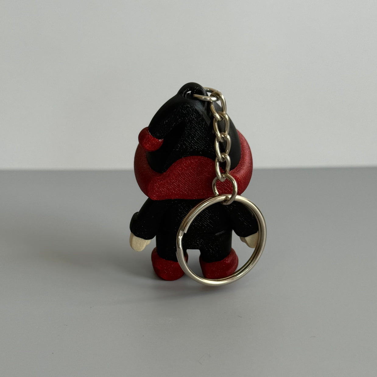 Back perspective of a small gnome keychain with a black and red hat with red shoes on a gray surface