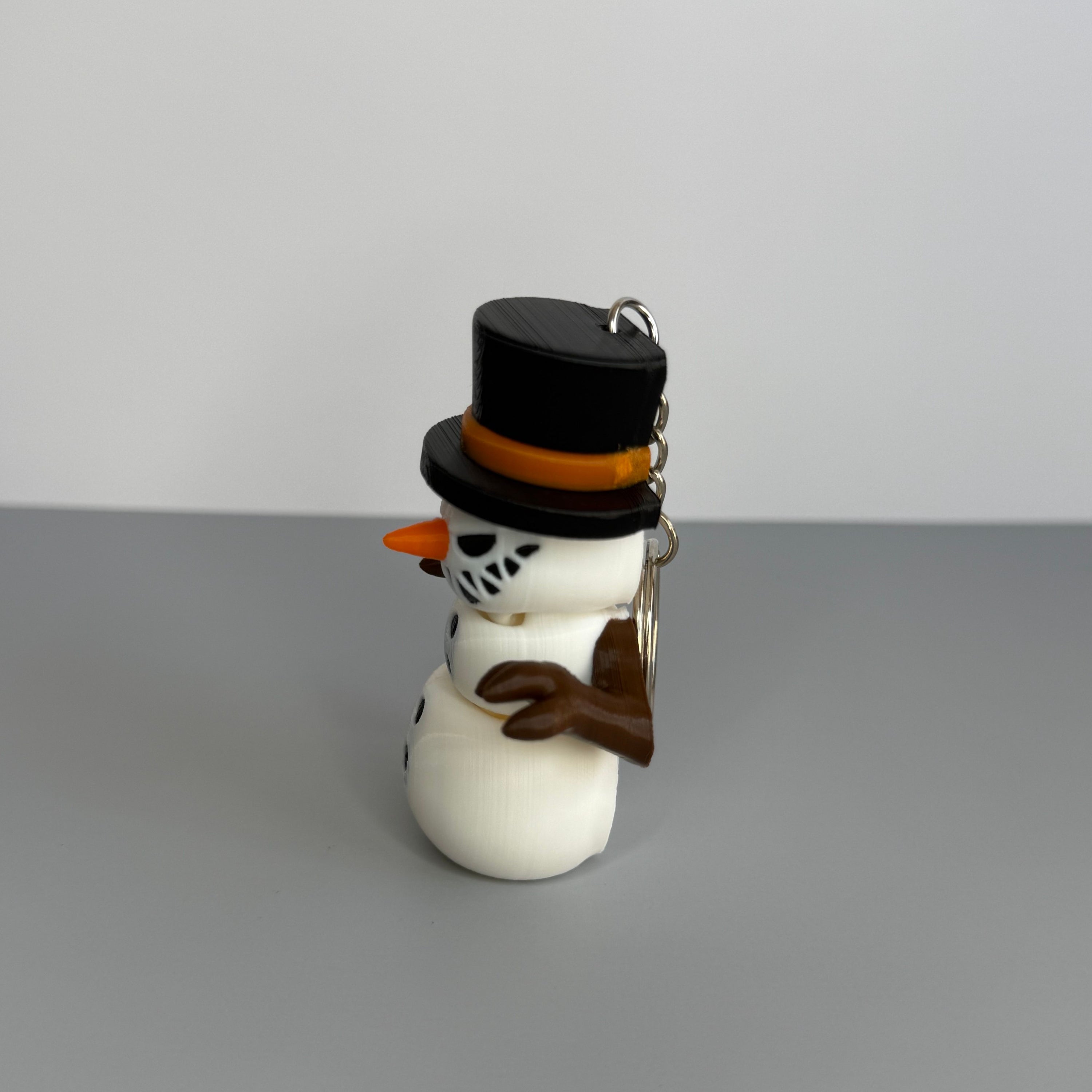 Right side of a snowman keychain with a black top hat, orange carrot nose, and sinister smile on a gray background.