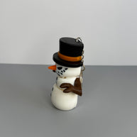 Right side of a snowman keychain with a black top hat, orange carrot nose, and sinister smile on a gray background.