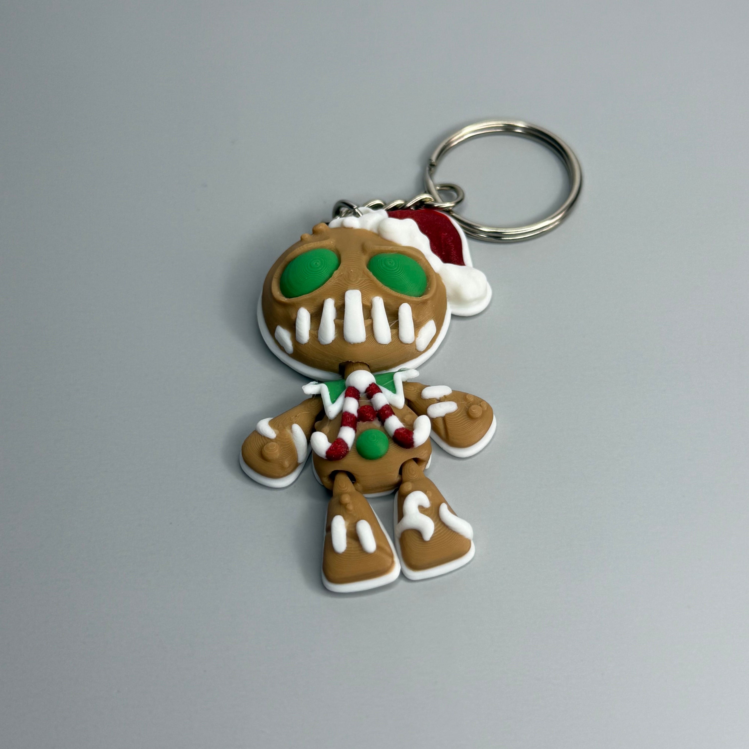Keychain shaped like a gingerbread man with green eyes, candy cane shaped bowtie, and a Santa hat on a gray background 