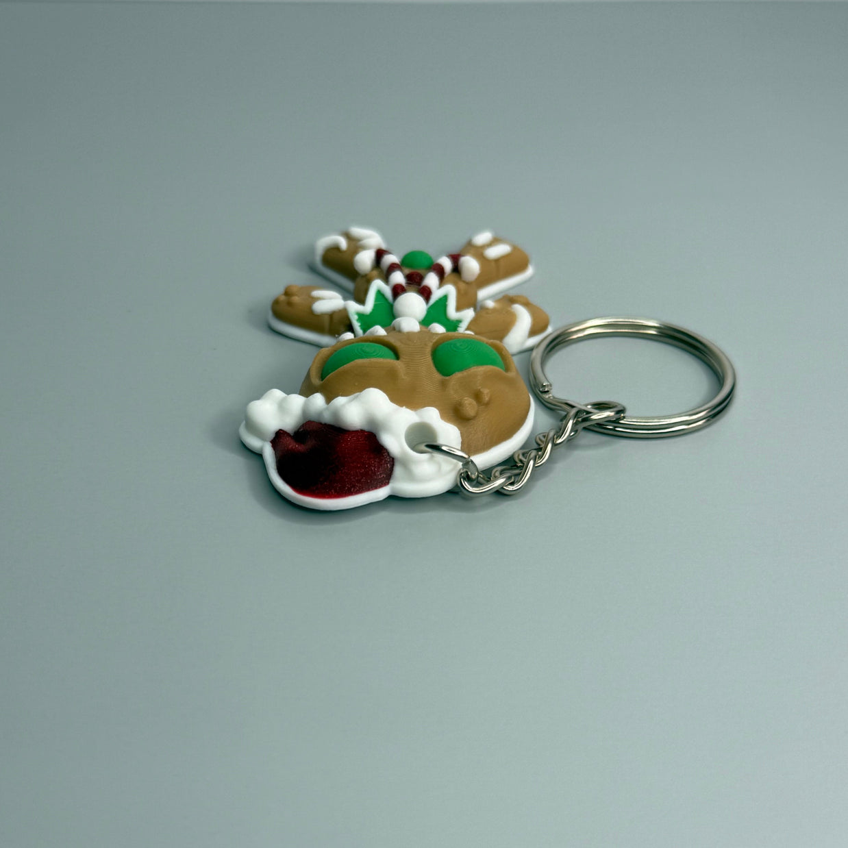 Keychain shaped like a gingerbread man with green eyes, candy cane shaped bowtie, and a Santa hat on a gray background 