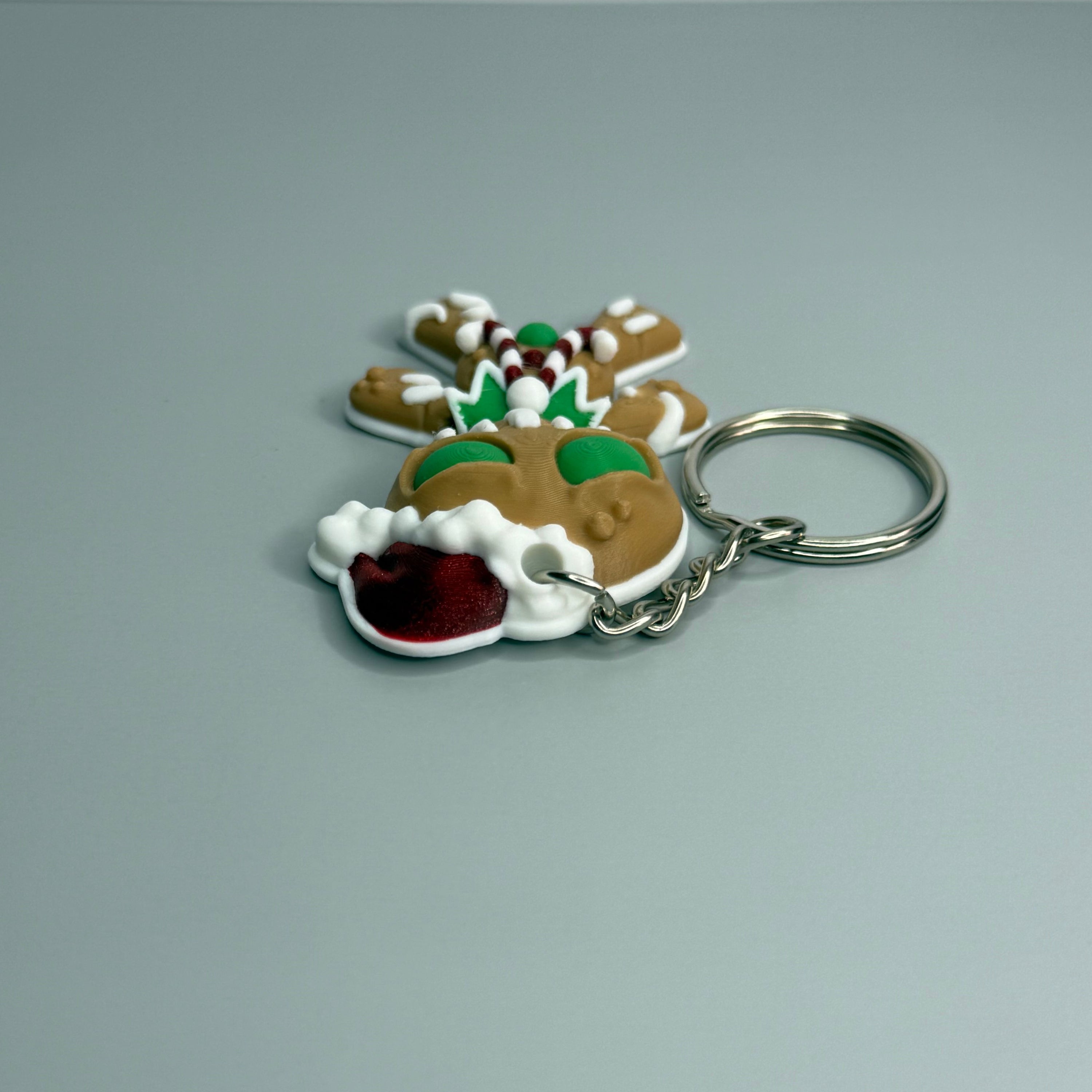 Keychain shaped like a gingerbread man with green eyes, candy cane shaped bowtie, and a Santa hat on a gray background 