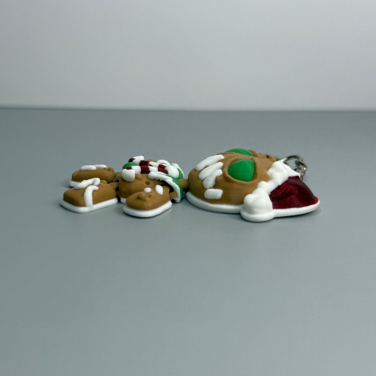 Right side of keychain shaped like a gingerbread man with green eyes, candy cane shaped bowtie, and a Santa hat on a gray background.