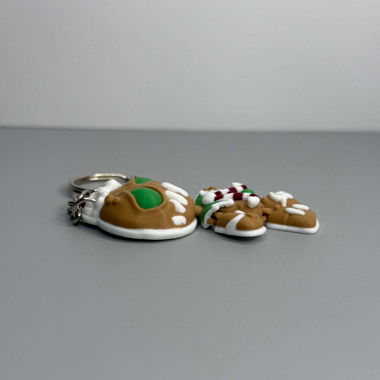 Left side of keychain shaped like a gingerbread man with green eyes, candy cane shaped bowtie, and a Santa hat on a gray background.