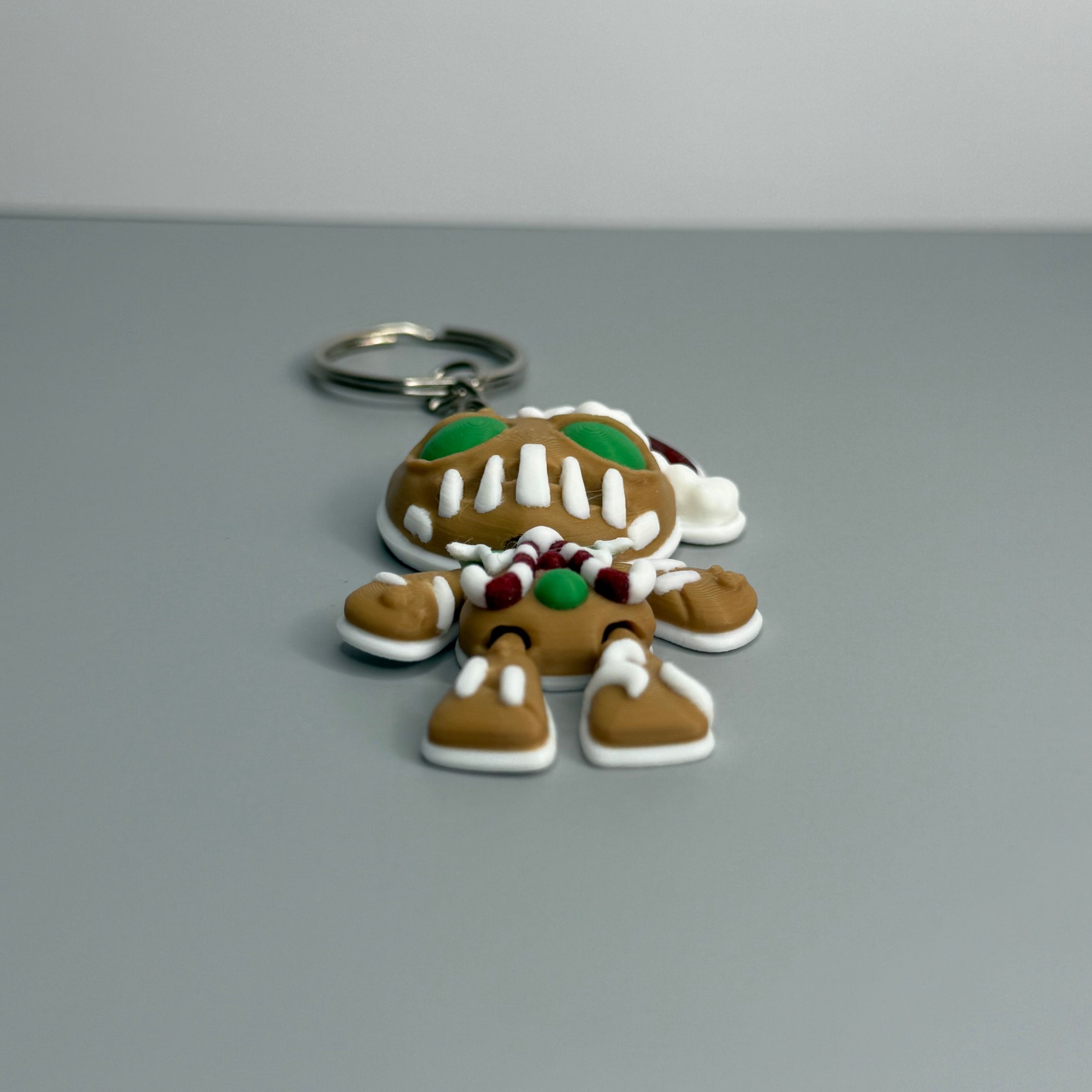 Keychain shaped like a gingerbread man with green eyes, candy cane shaped bowtie, and a Santa hat on a gray background.