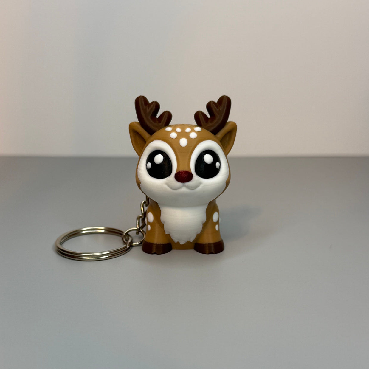 Front perspective of reindeer-shaped keychain with a red, shimmer nose on a gray background. 