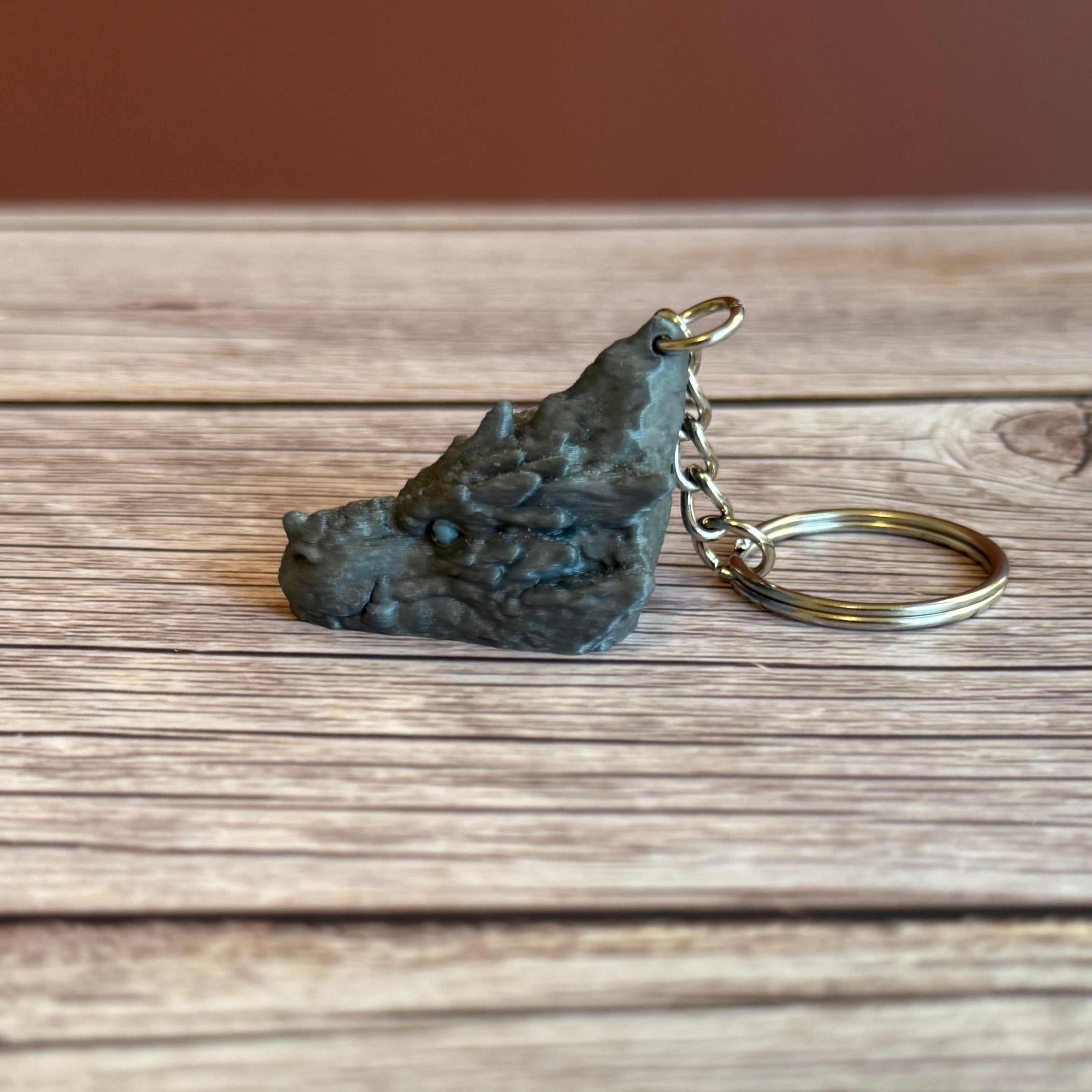Side of a 3D printed gray dragon head keychain with blue eyes on a wooden surface