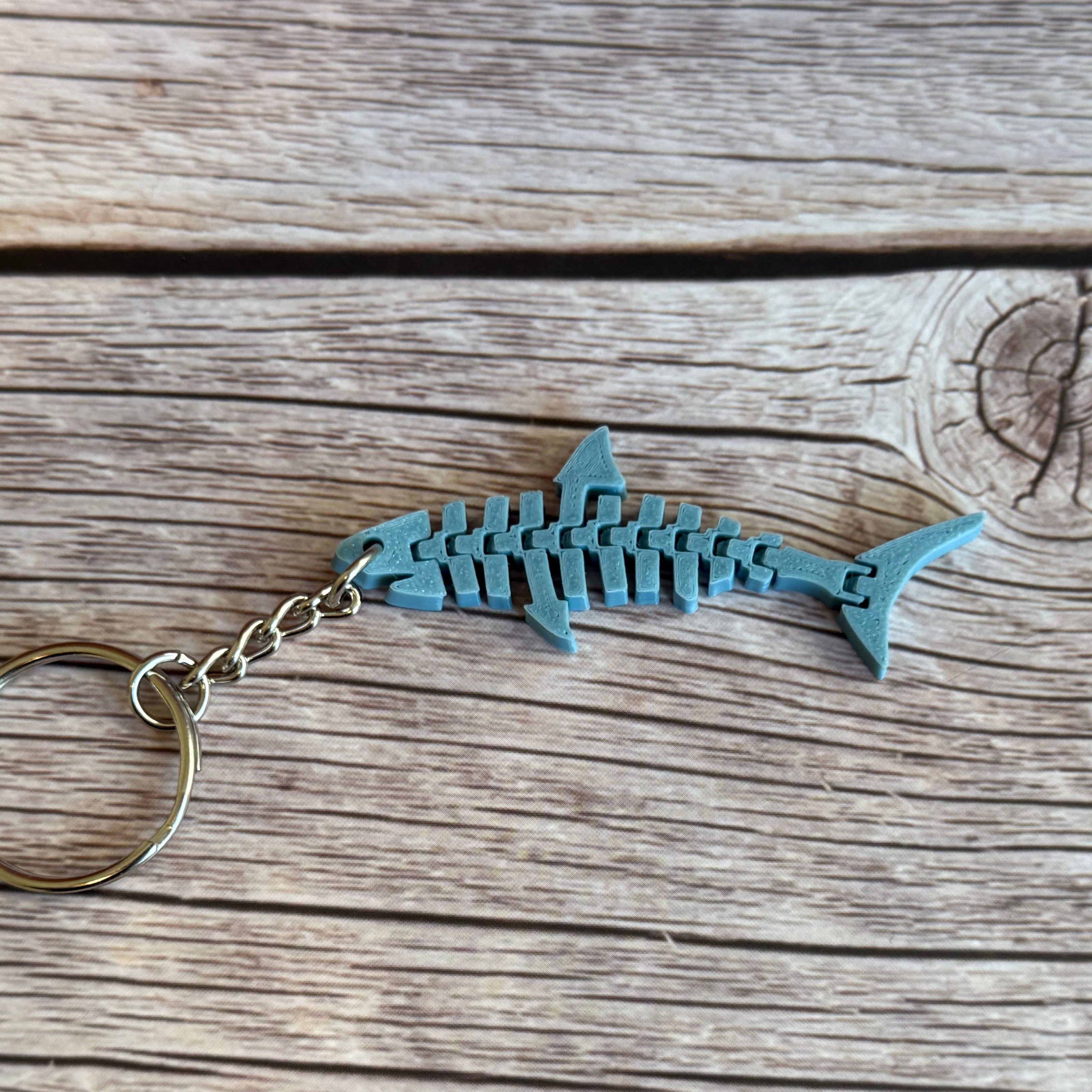 3D printed blue-gray flat flexi shark keychain on a wooden background.