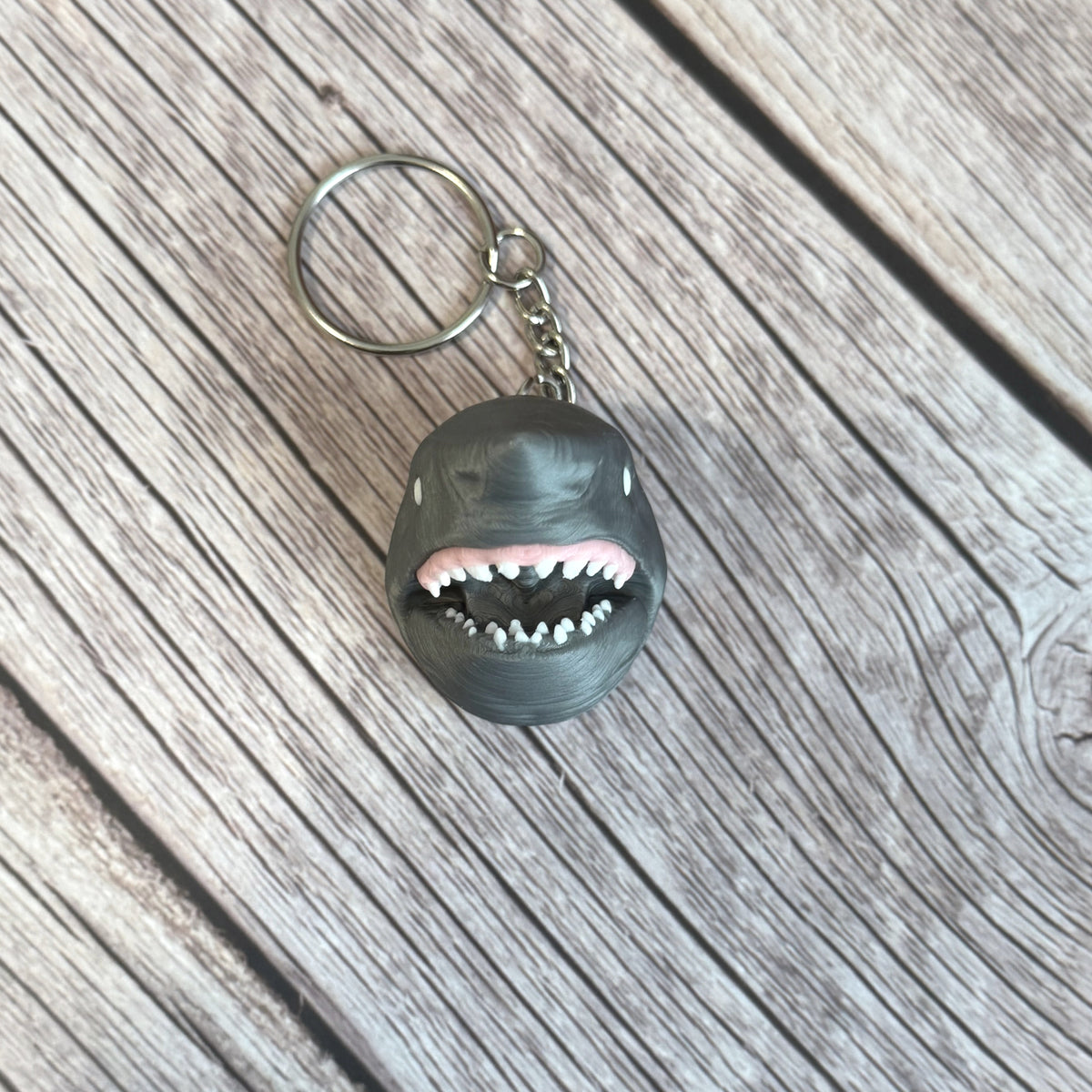 3D printed gray keychain in the shape of a shark head with open mouth, set on a wooden surface