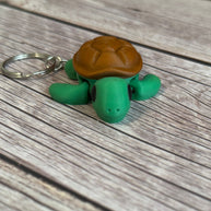 3D printed turtle-shaped keychain with brown shell and green body on a wooden surface