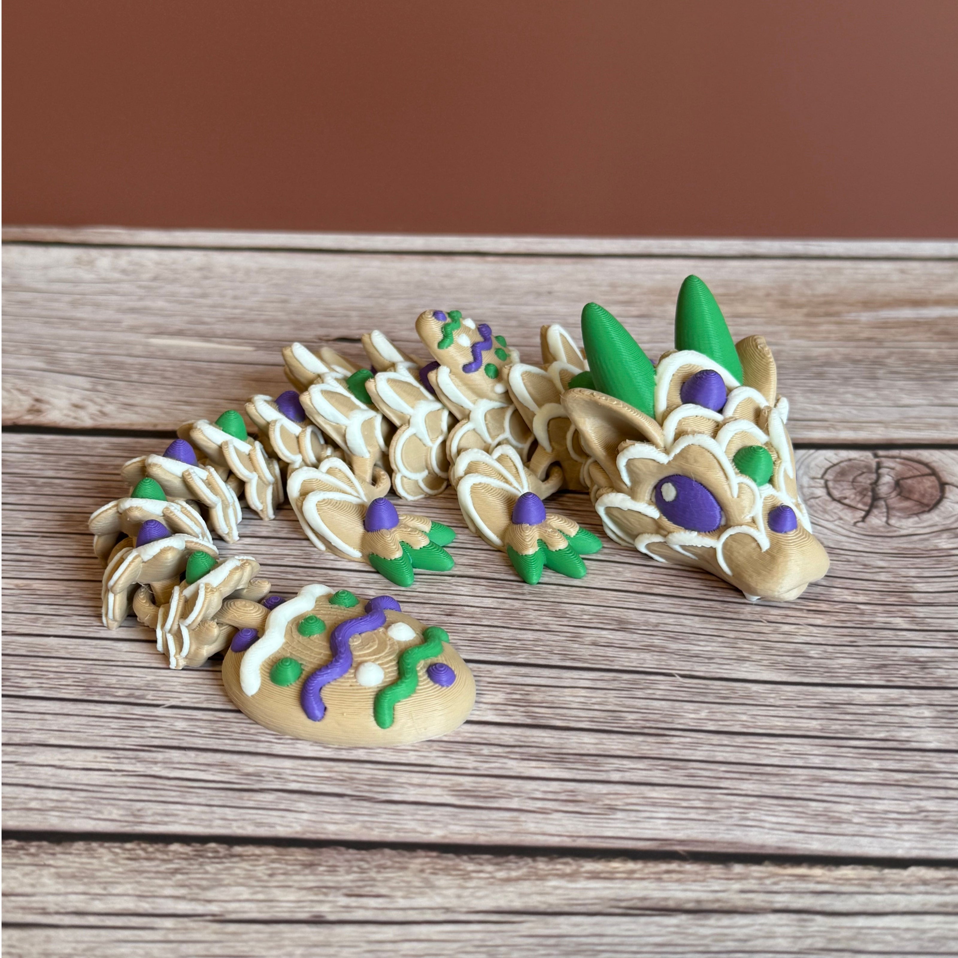 3D printed articulated dragon figure resembling a cookie with beige, green, purple, and white colors. Placed on a wooden surface