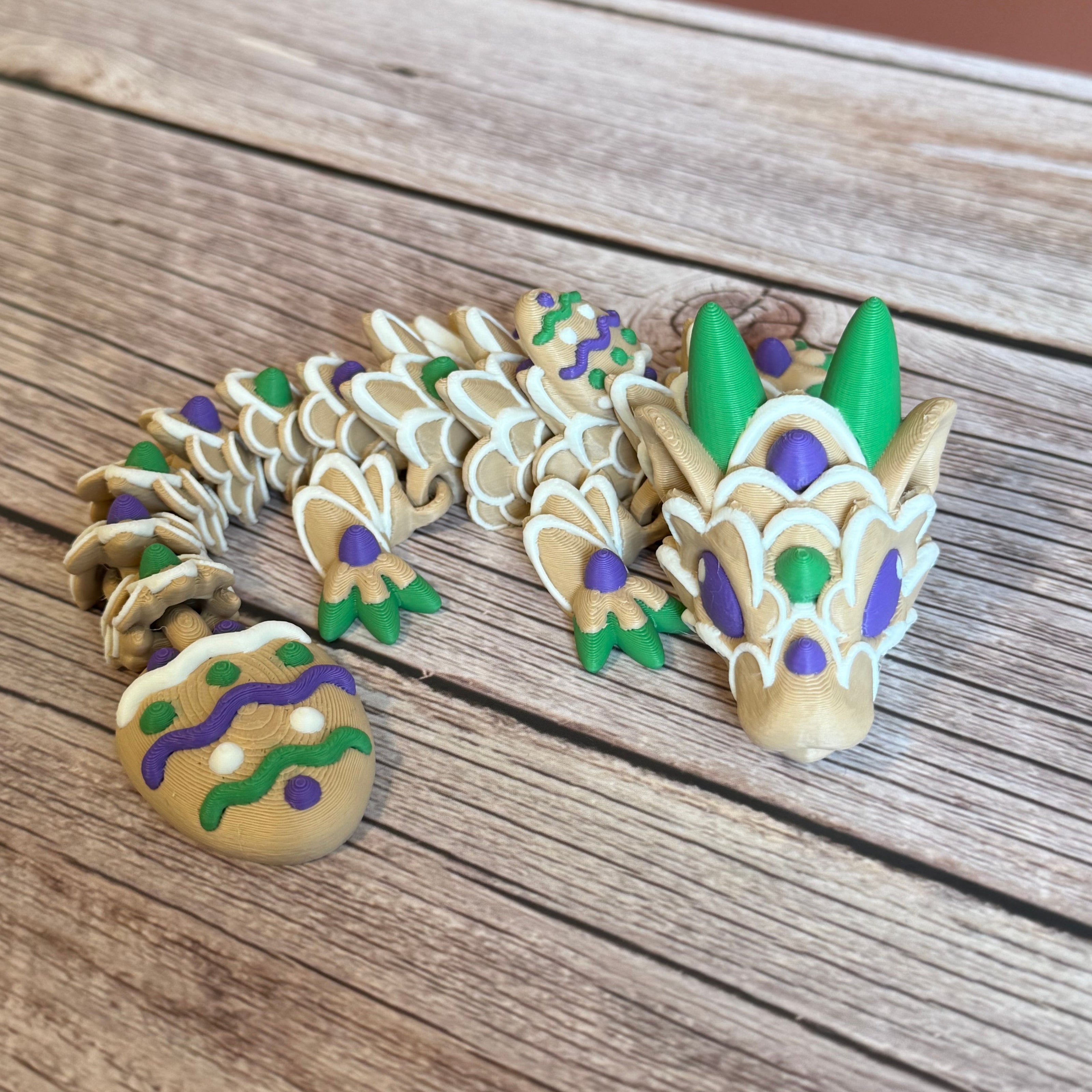 3D printed articulated dragon figure resembling a cookie with beige, green, purple, and white colors. Placed on a wooden surface