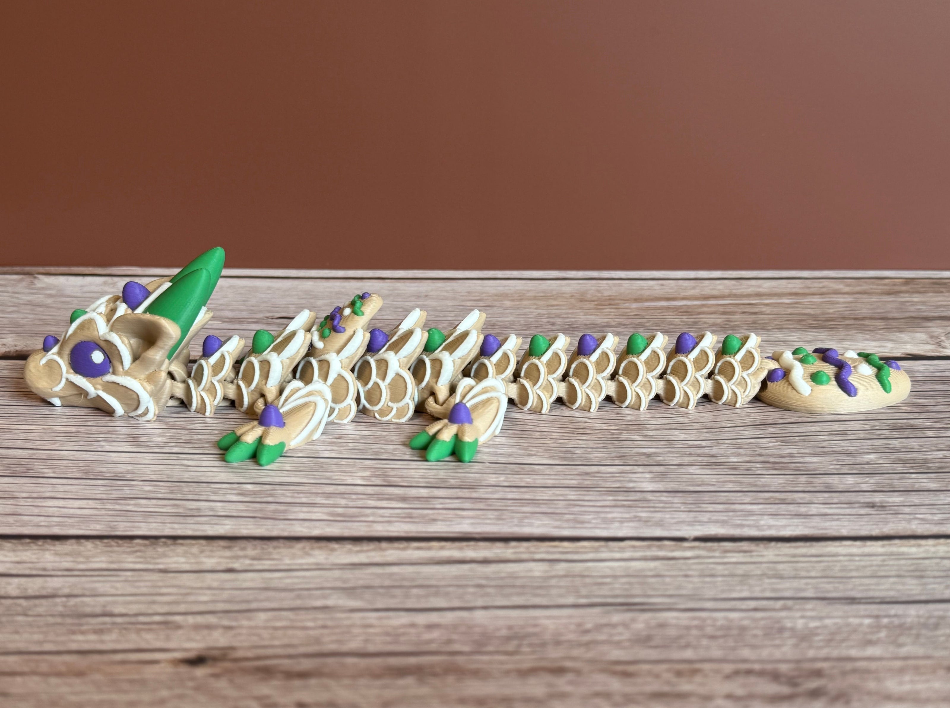Side of a 3D printed articulated dragon figure resembling a cookie with beige, green, purple, and white colors. Placed on a wooden surface
