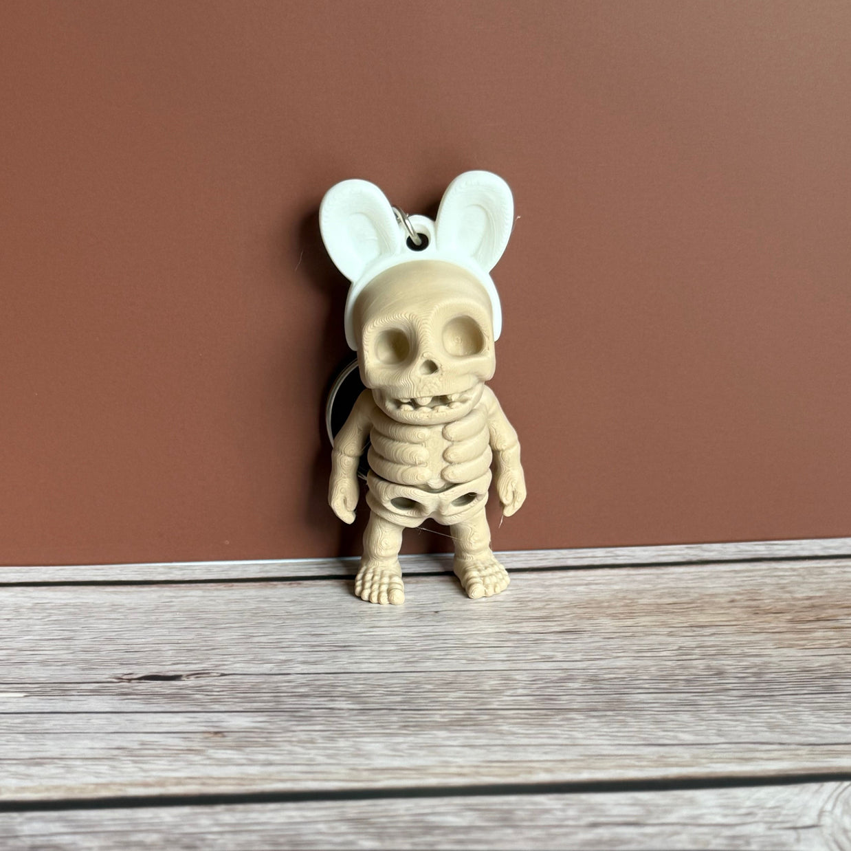 3D printed keychain of a beige skeleton with white bunny ears on a wooden surface, against a brown background