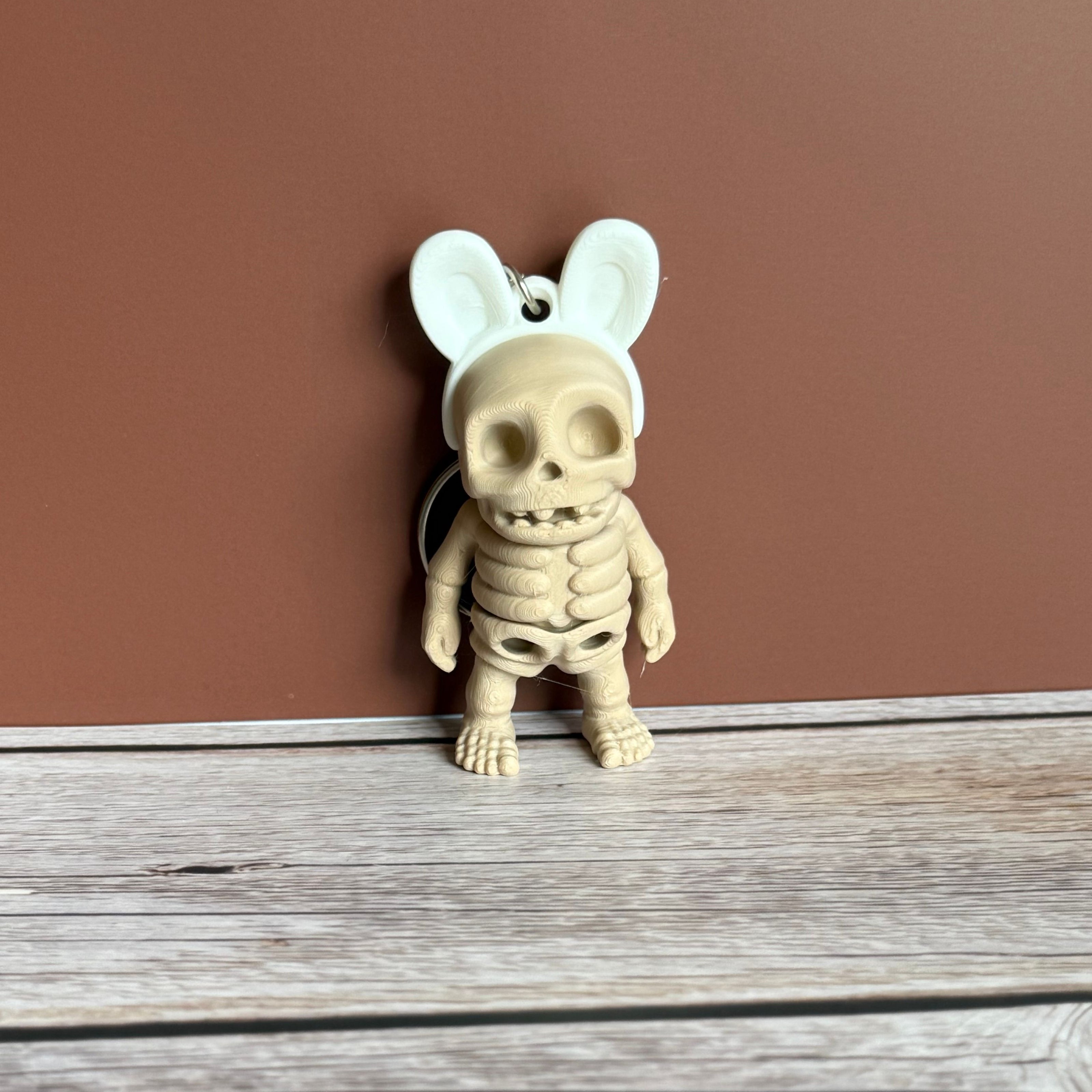 3D printed keychain of a beige skeleton with white bunny ears on a wooden surface, against a brown background