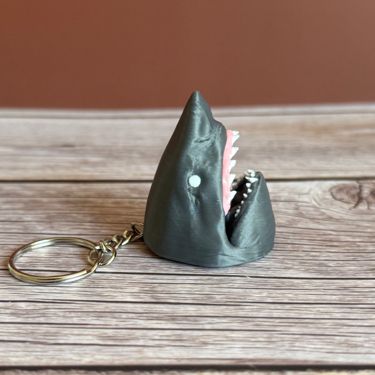 3D printed gray keychain in the shape of a shark head with open mouth, set against a wooden background.