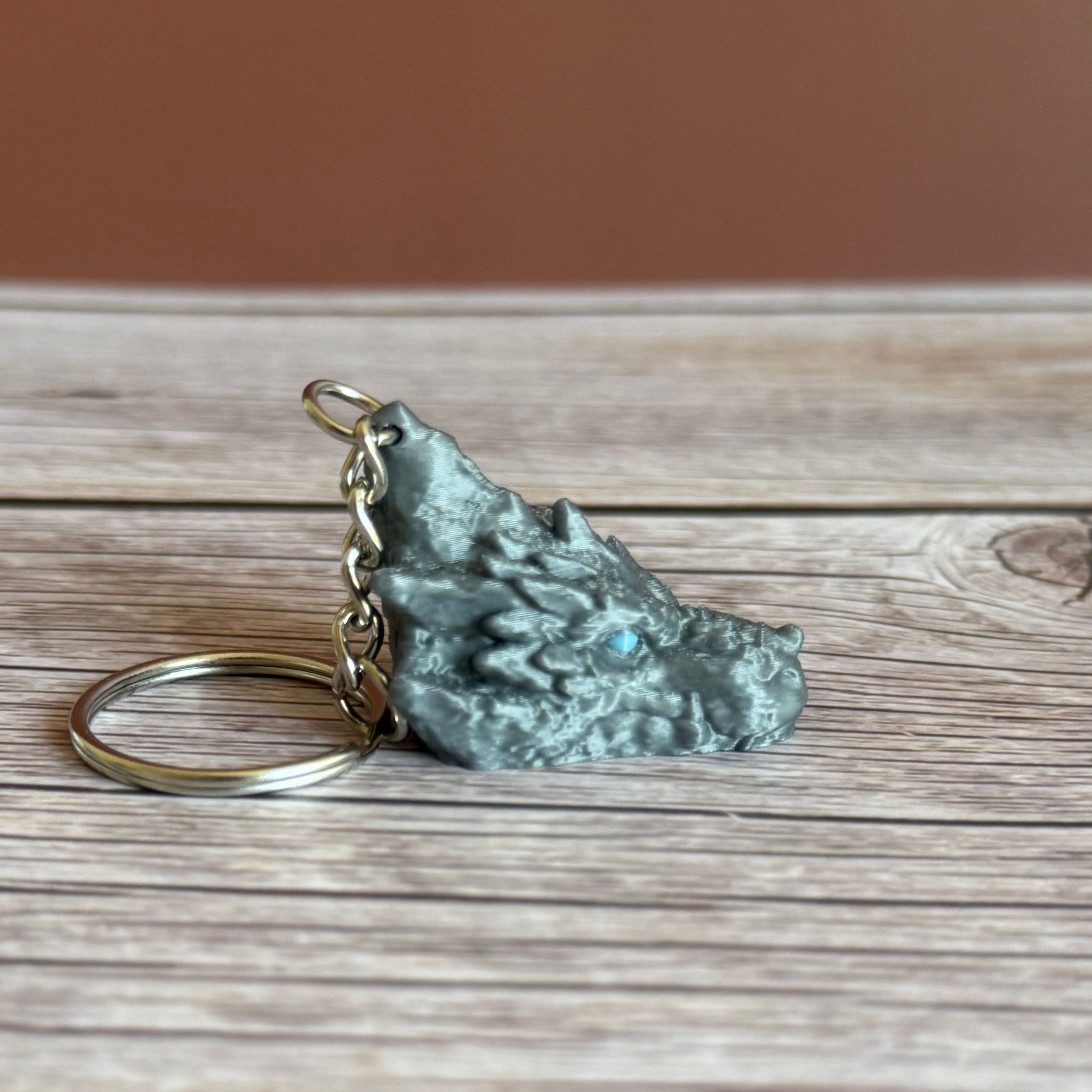 Side of a 3D printed gray dragon head keychain with blue eyes on a wooden surface