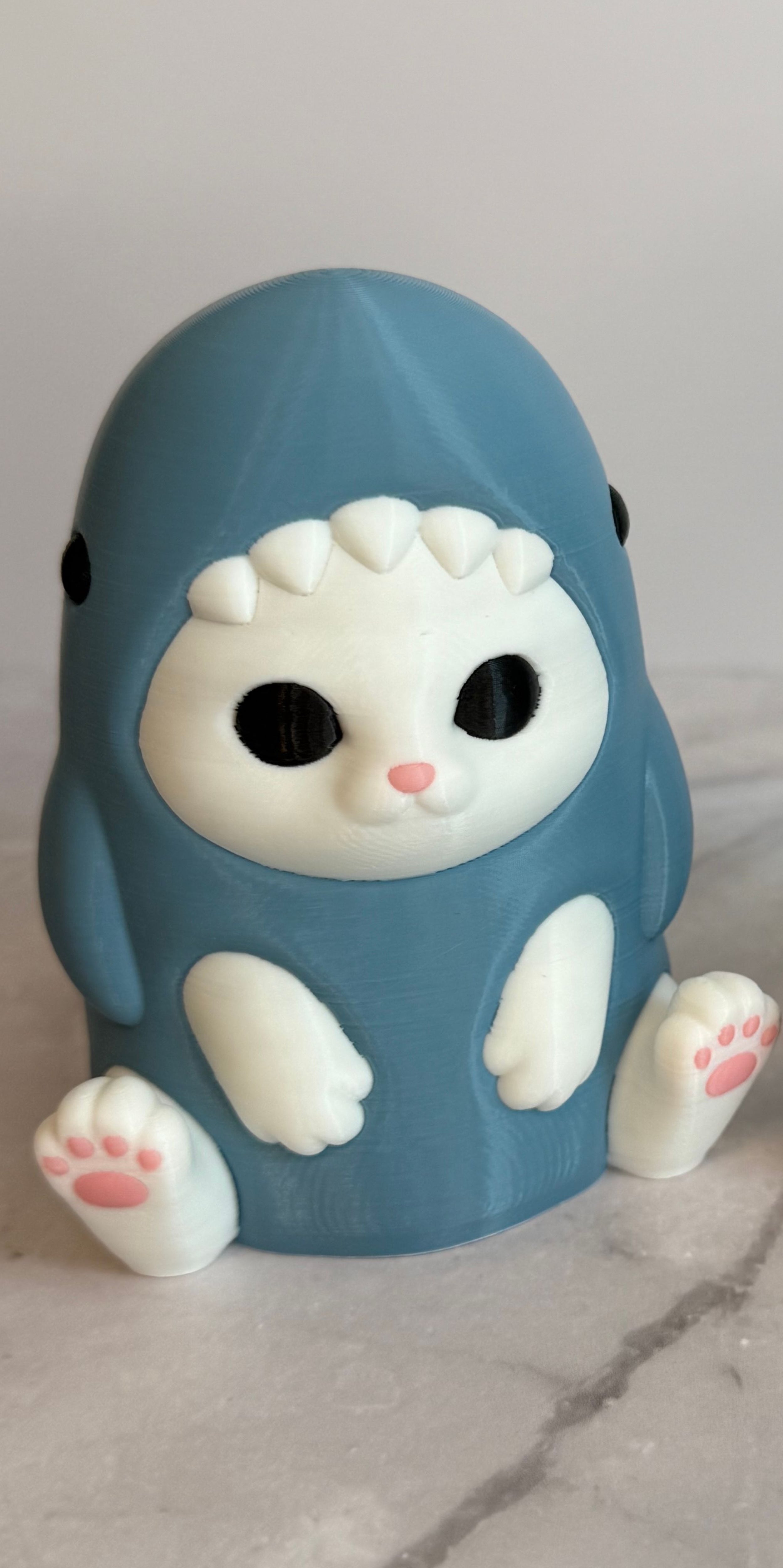3D printed figurine of a white cat wearing a shark costume in blue-gray color, placed on a marble surface