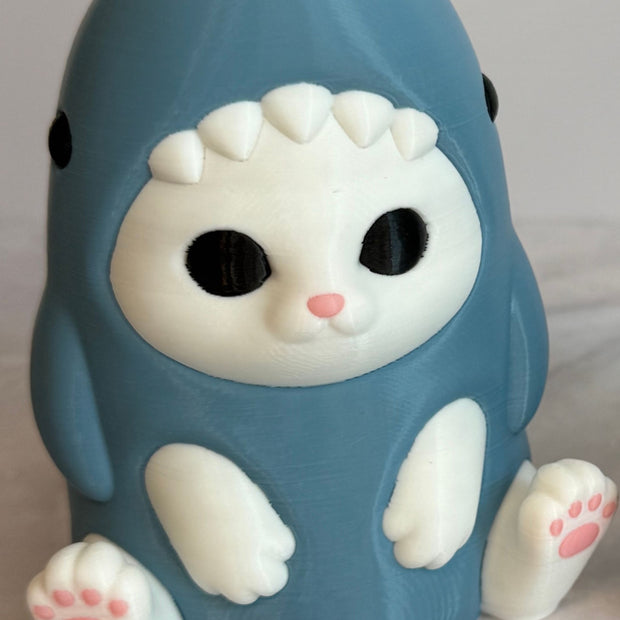 3D printed figurine of a white cat wearing a shark costume in blue-gray color, placed on a marble surface