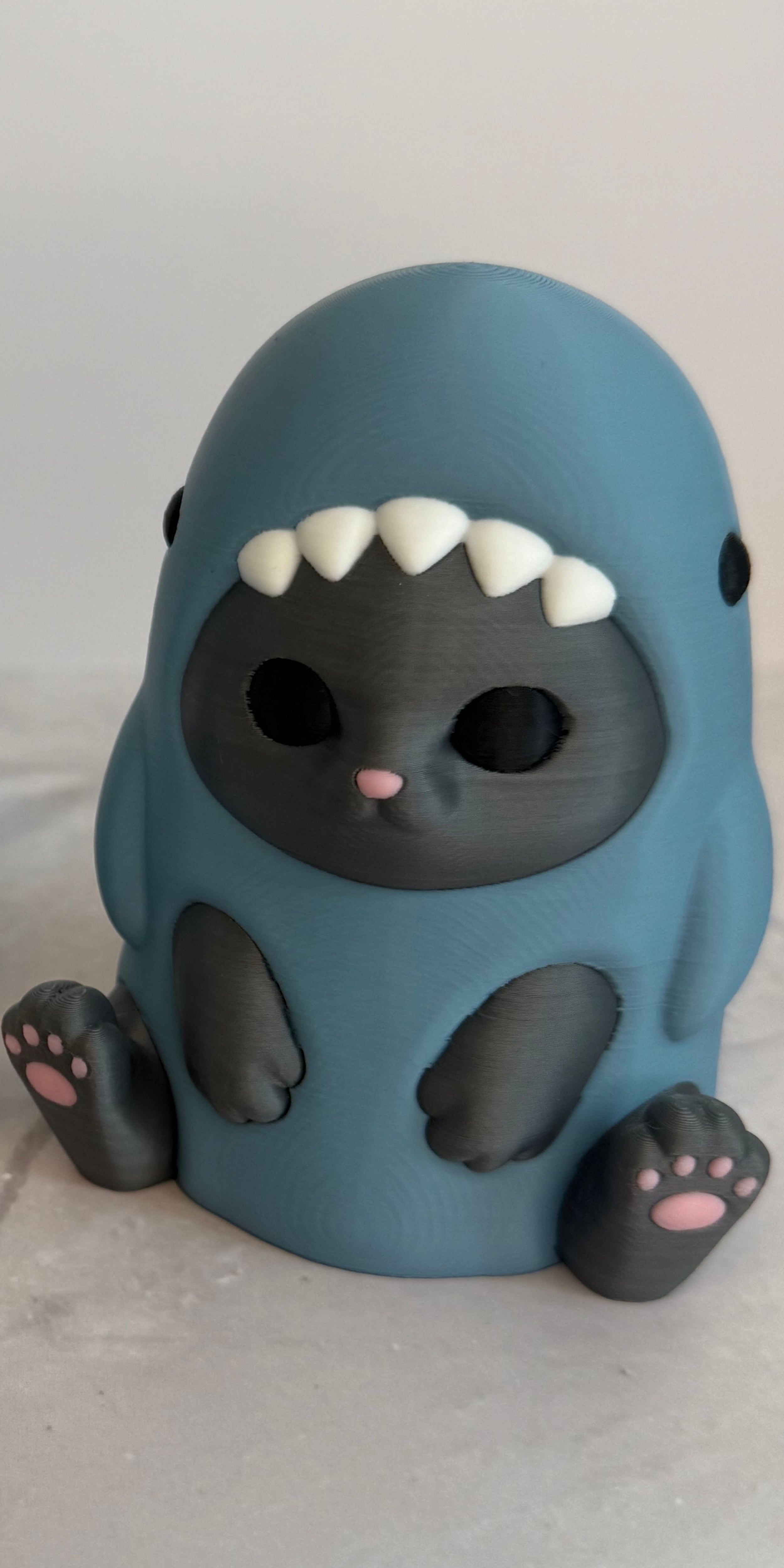 3D printed figurine of a gray cat wearing a shark costume in blue-gray color, placed on a marble surface