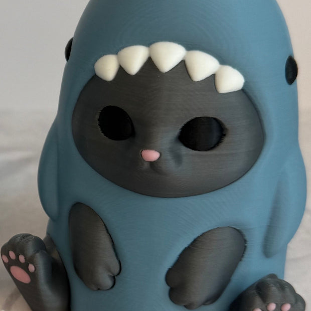 3D printed figurine of a gray cat wearing a shark costume in blue-gray color, placed on a marble surface