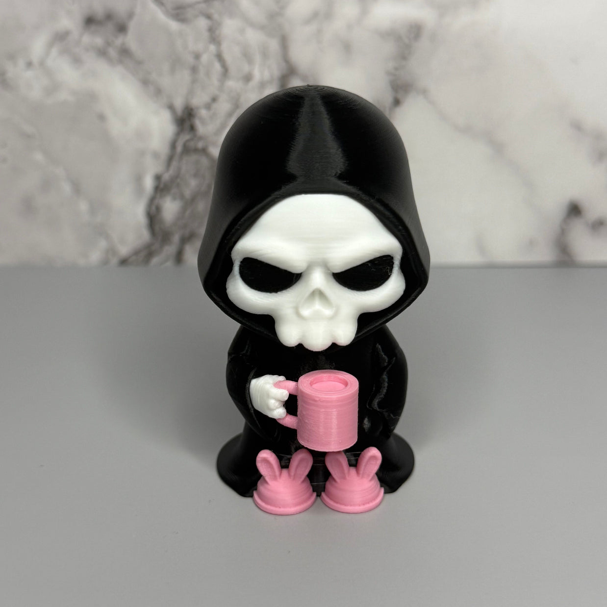 Skull figure in a black hood holding a pink mug on a gray surface with marble background