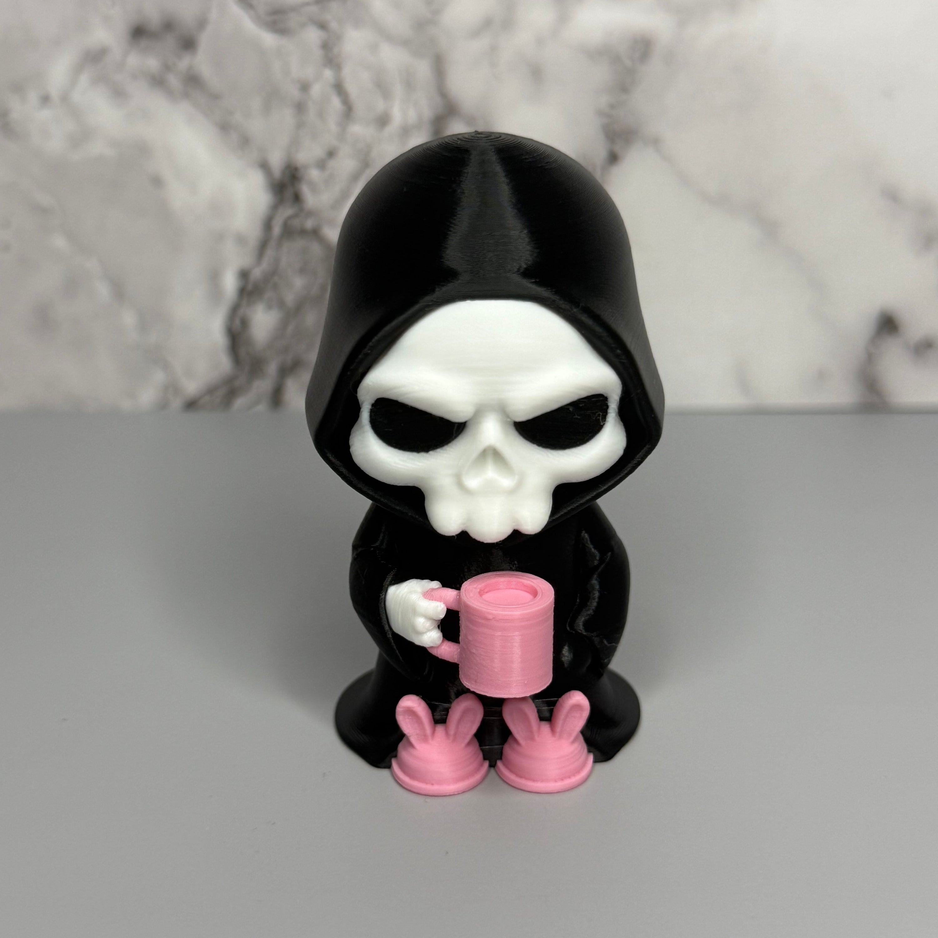 Skull figure in a black hood holding a pink mug on a gray surface with marble background