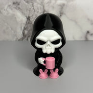 Skull figure in a black hood holding a pink mug on a gray surface with marble background