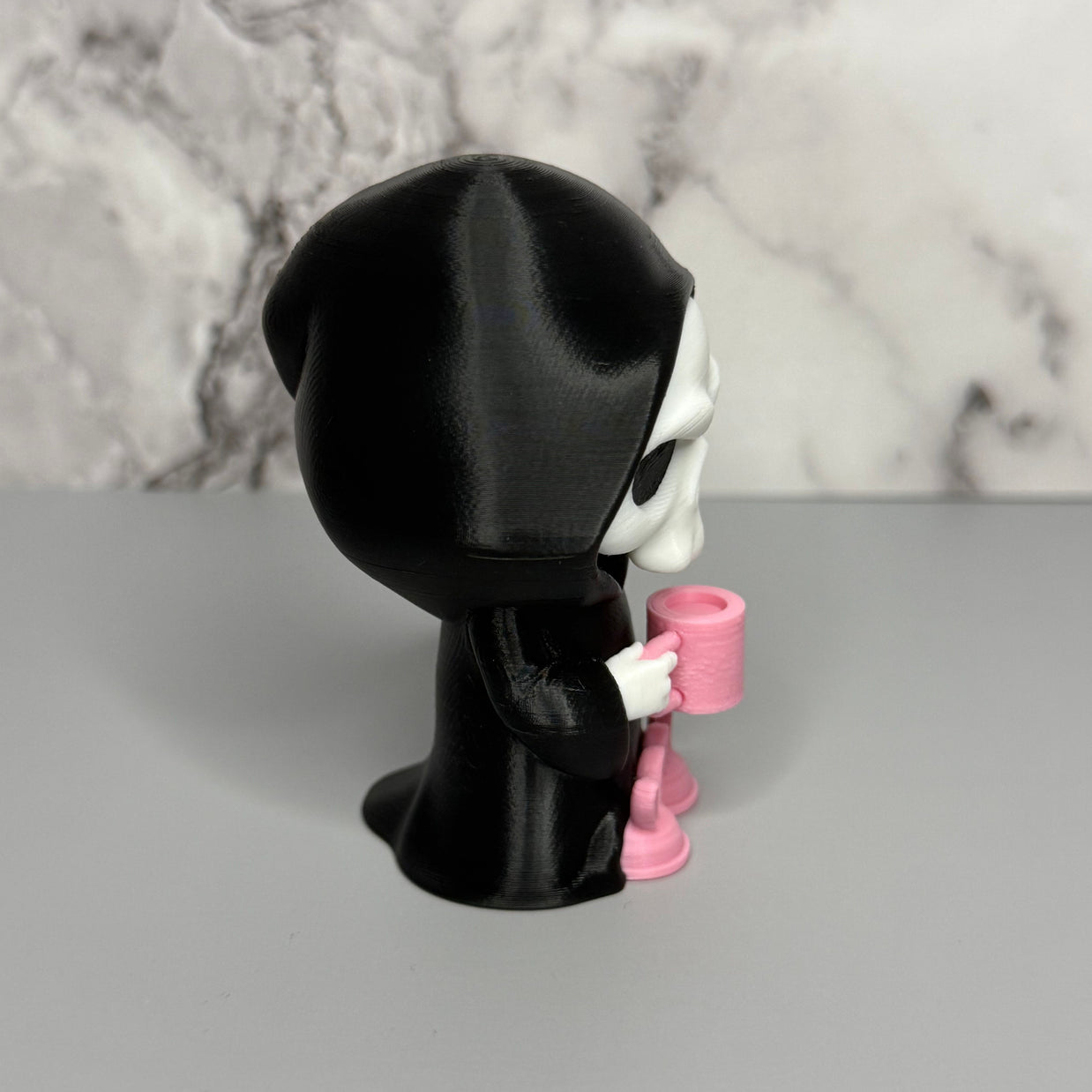 Left side of skull figure in a black hood holding a pink mug on a gray surface with marble background