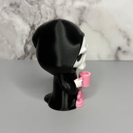 Left side of skull figure in a black hood holding a pink mug on a gray surface with marble background