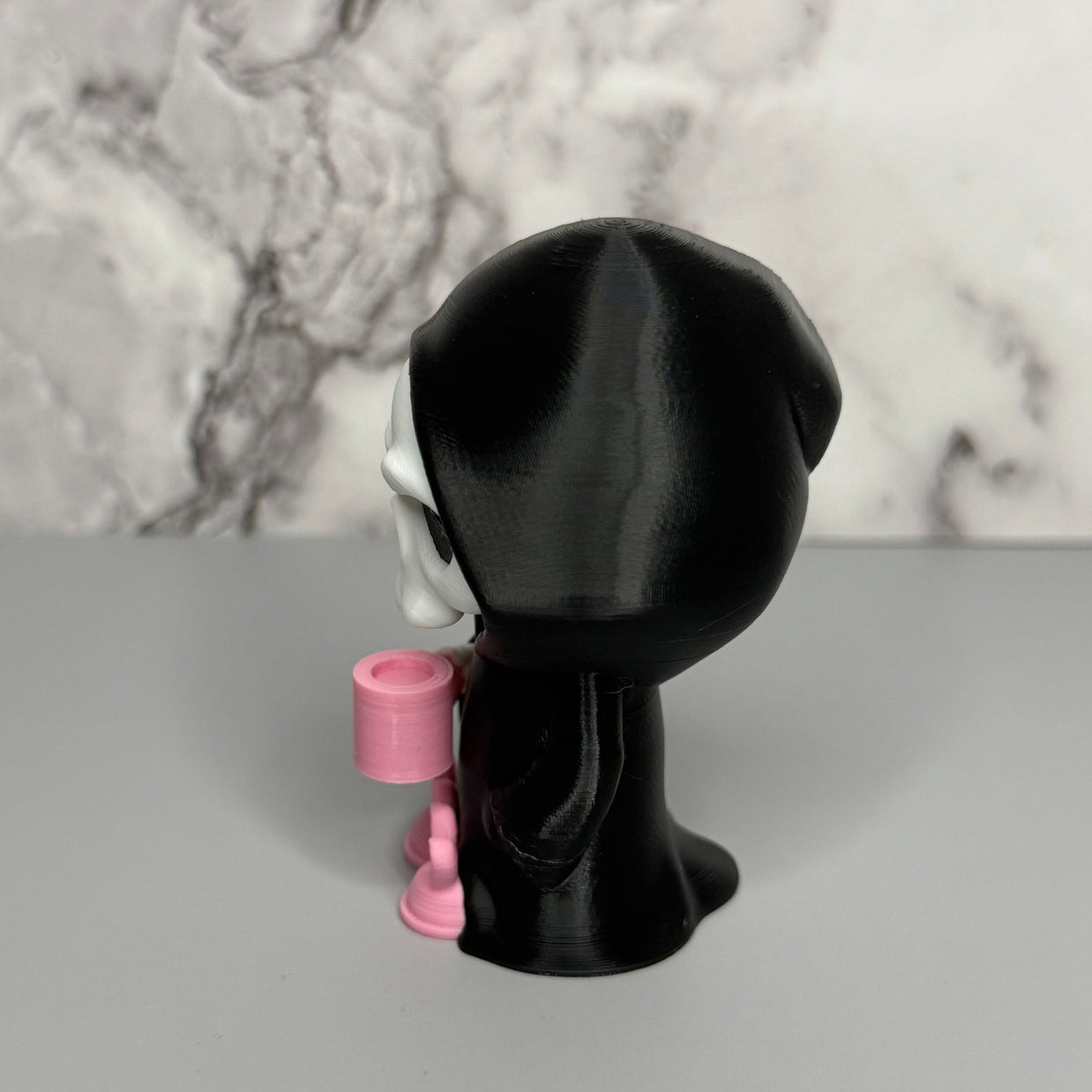 Right side of skull figure in a black hood holding a pink mug on a gray surface with marble background