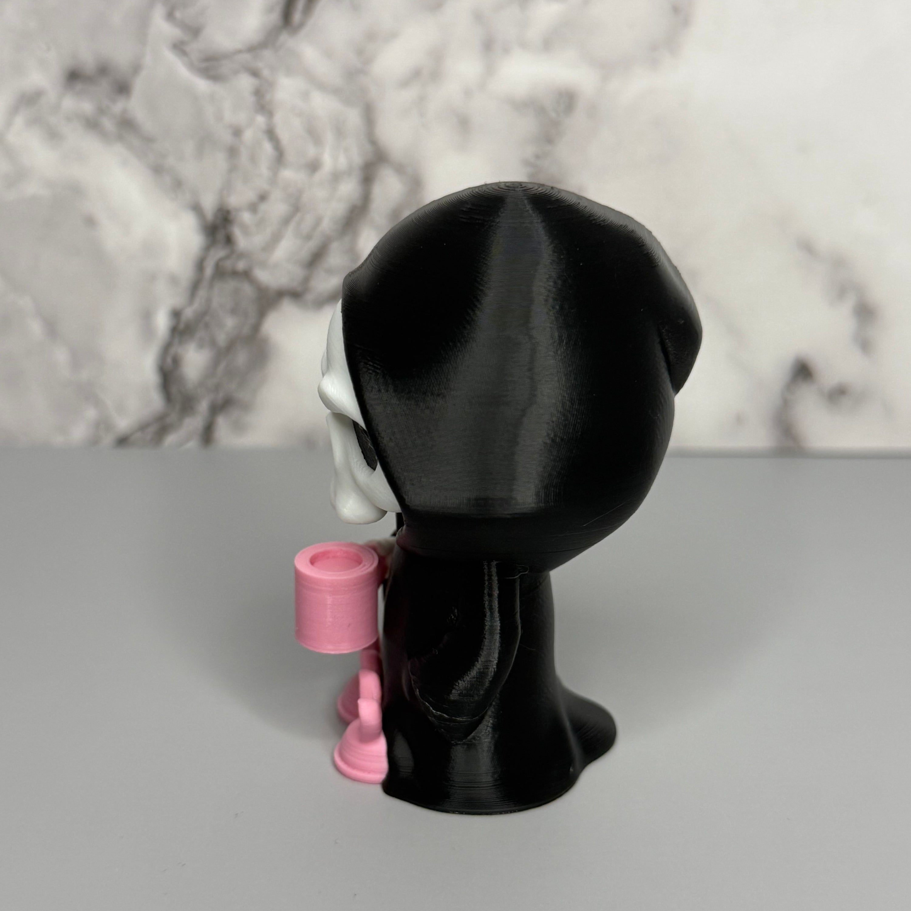 Right side of skull figure in a black hood holding a pink mug on a gray surface with marble background