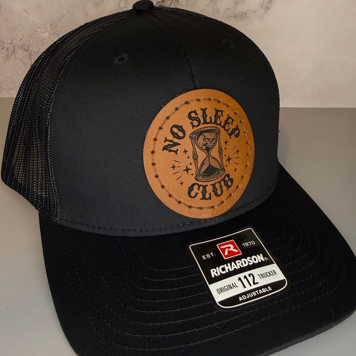 Black cap with a Richardson sticker and a brown leather patch featuring an hourglass and skull design with the phrase 