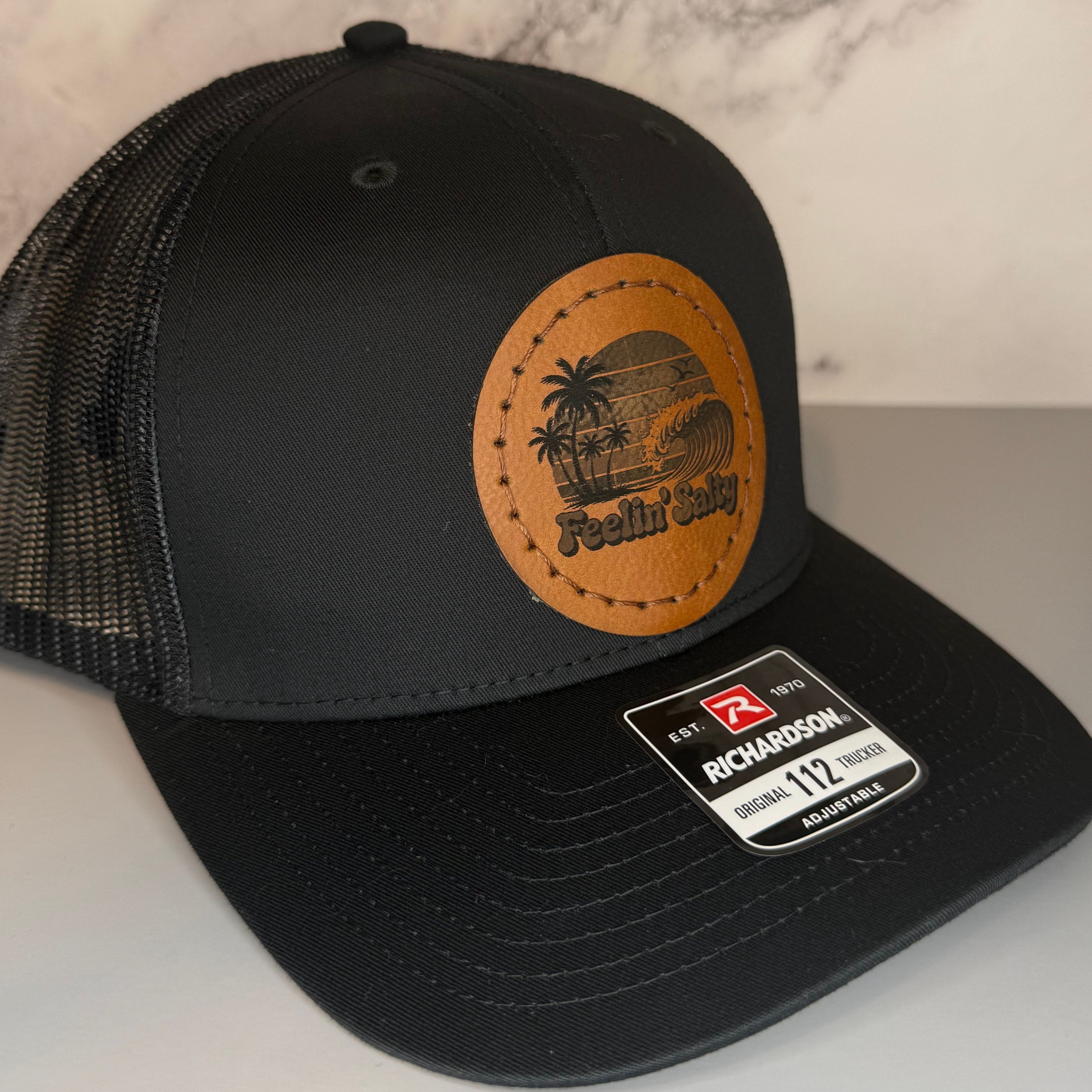 Black cap with a Richardson sticker and a brown leather patch featuring a palm tree and ocean wave design with the phrase 