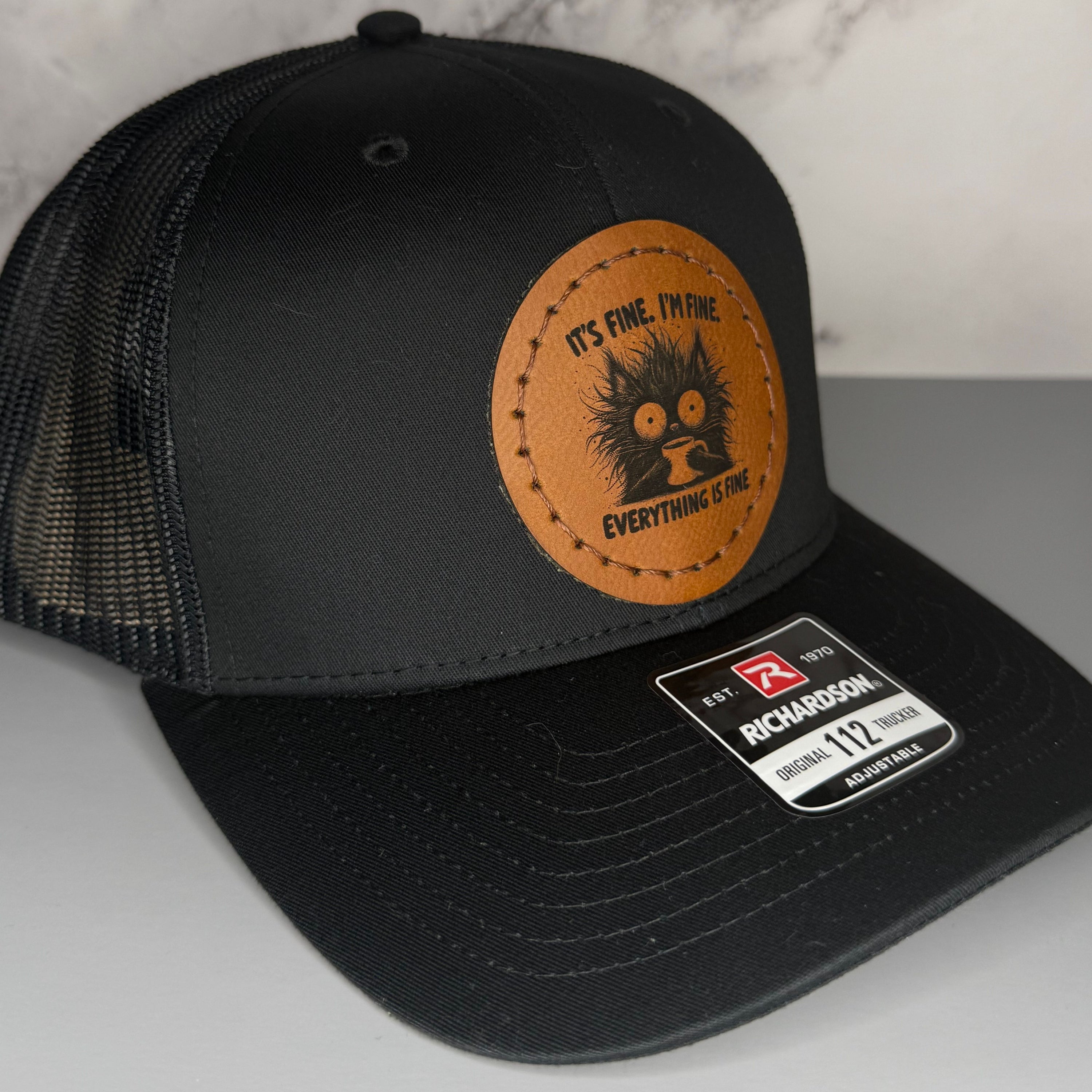 Black cap with a Richardson sticker and a brown leather patch featuring a cat and coffee design with the phrase 