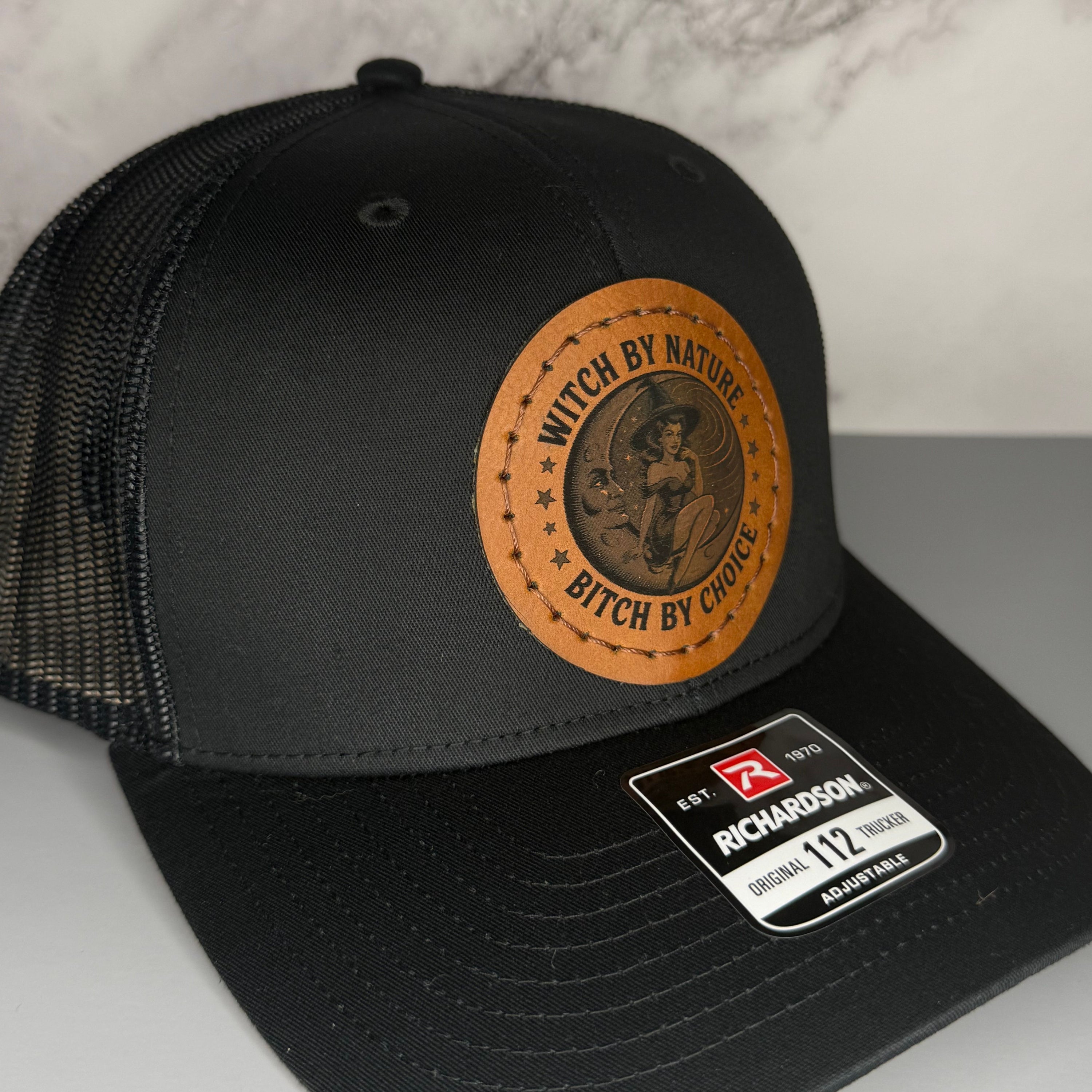 Black cap with a Richardson sticker and a brown leather patch featuring a witch and moon design with the phrase 