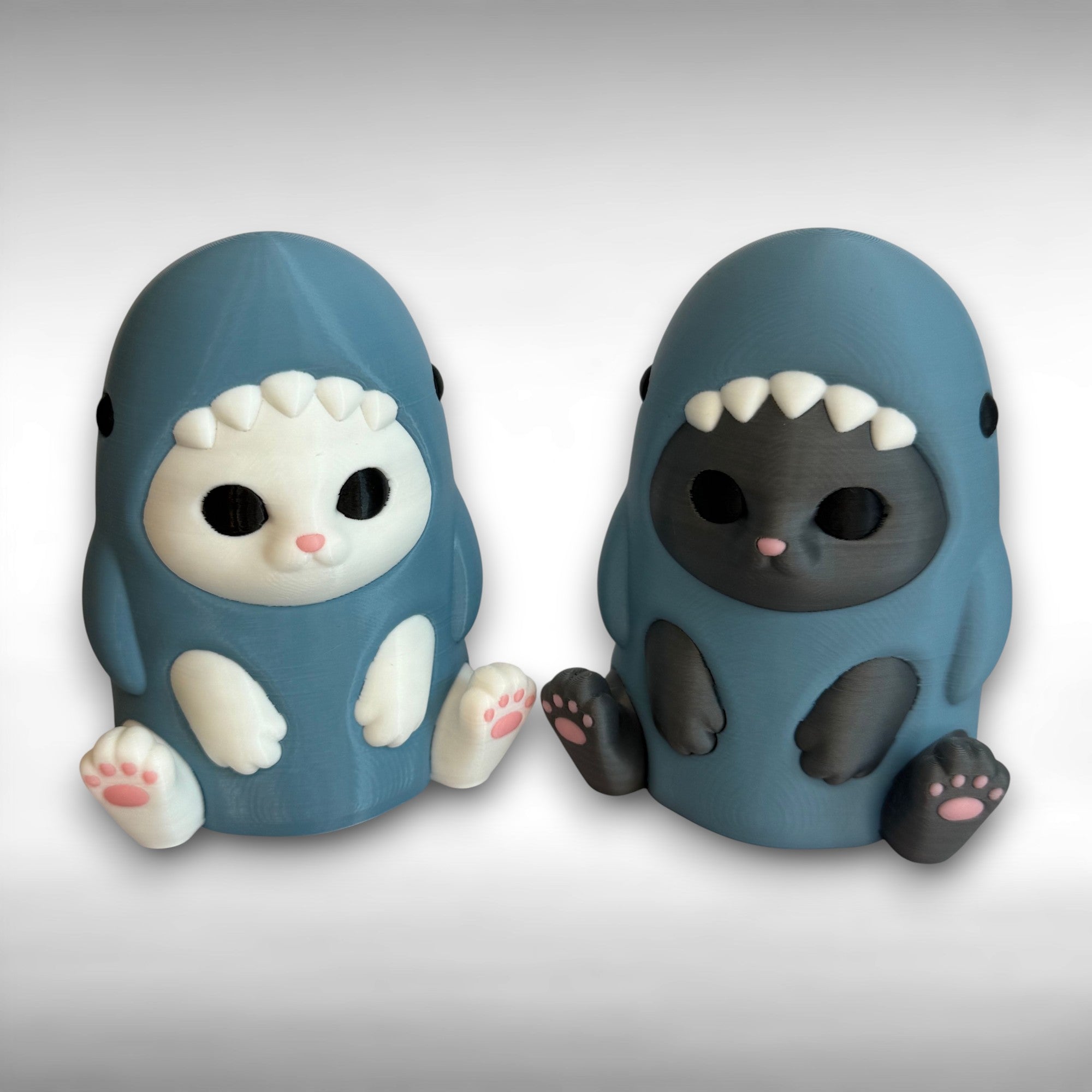 Two 3D printed cat figurines dressed in blue-gray shark-appearing costumes, one white cat and one gray cat, against a white background