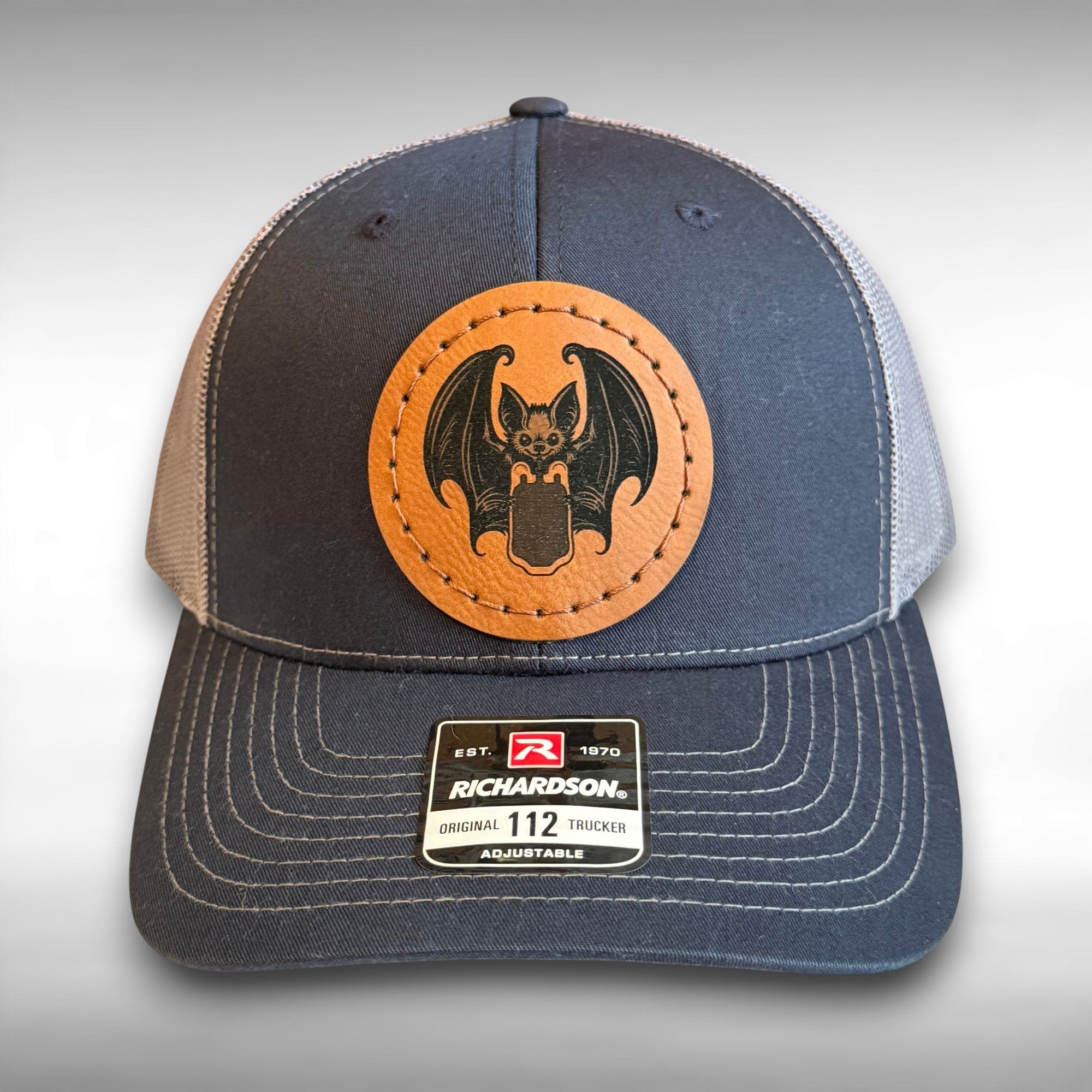 Blue and gray cap with a circular leather patch featuring a bat drinking from a blood bag design on a gray background