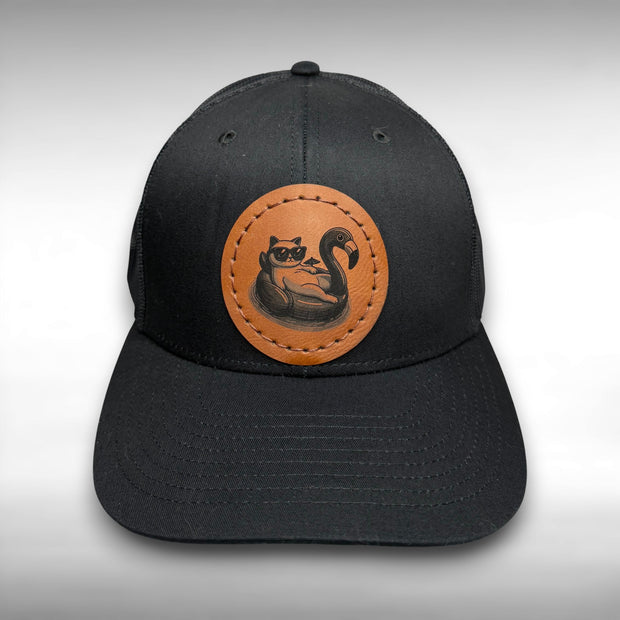 Black cap with a brown leather patch featuring a cat with sunglasses on a flamingo pool float on a gray background