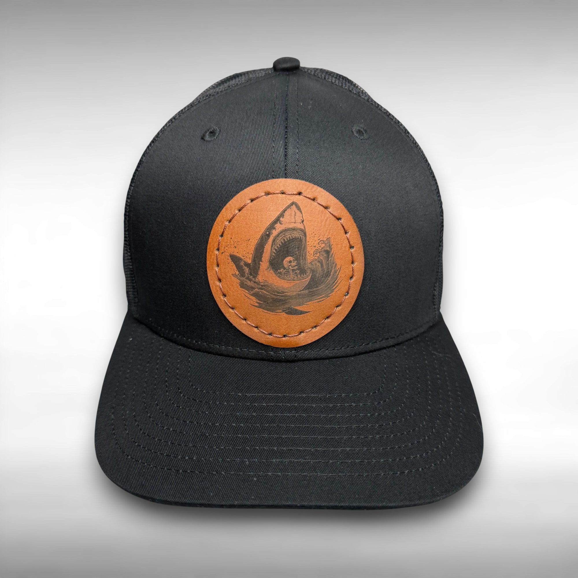 Black trucker hat with a brown leather patch featuring a shark and skeleton design on a gray background