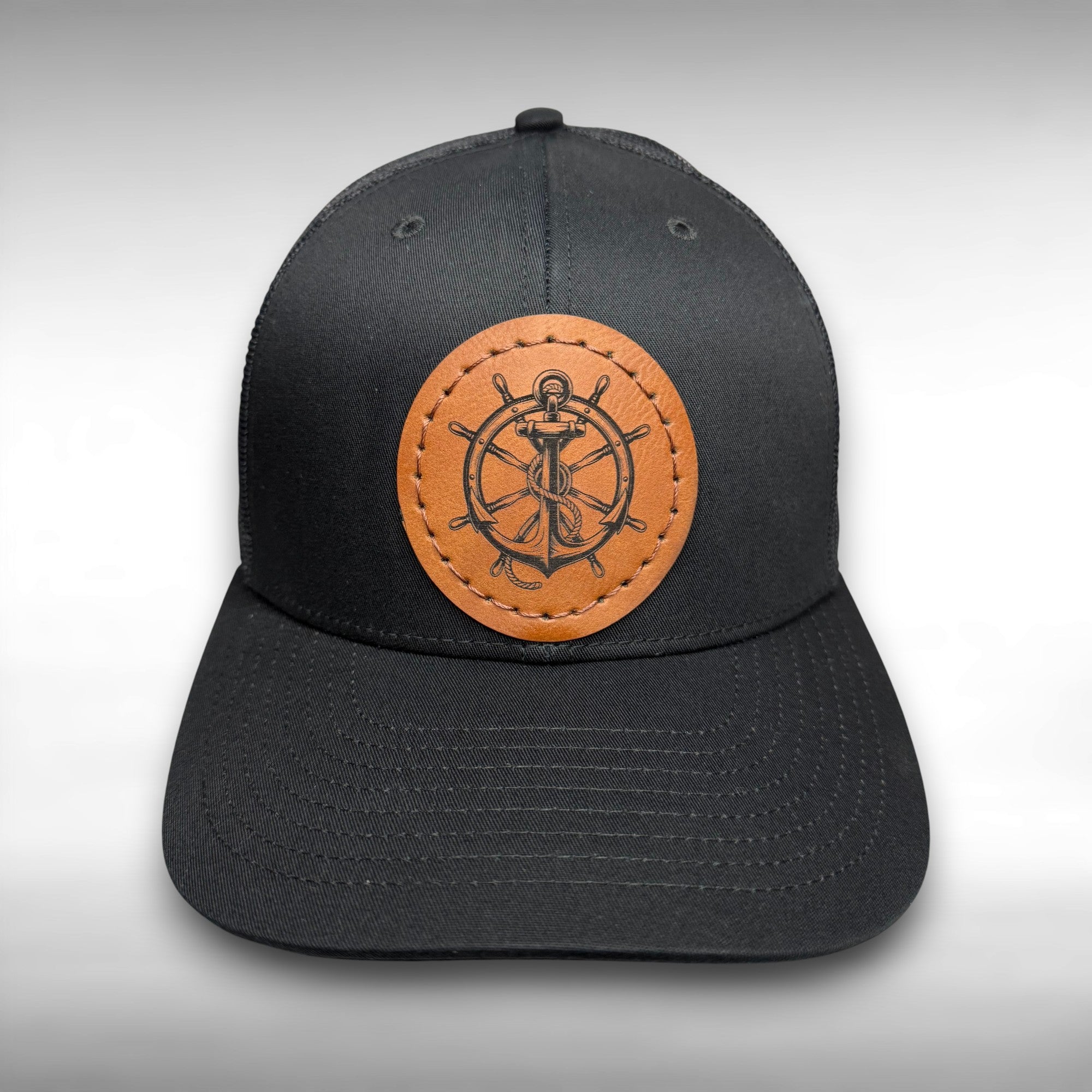 Black cap with a brown leather patch featuring a ship's wheel and anchor design on a gray background