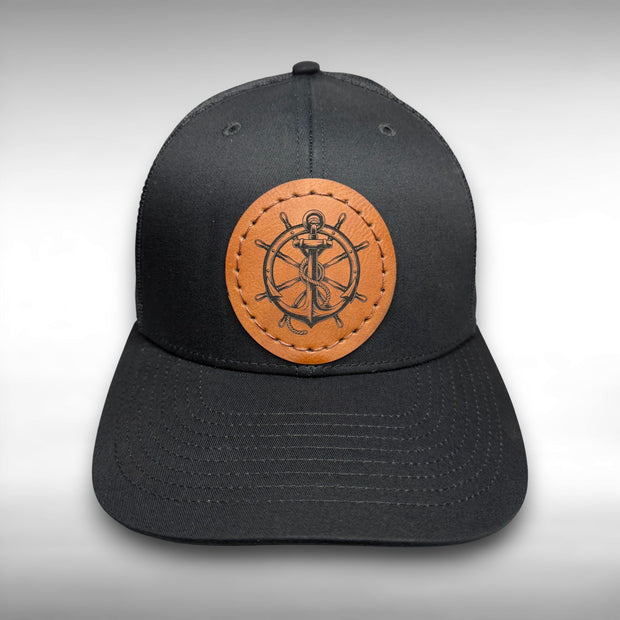 Black cap with a brown leather patch featuring a ship's wheel and anchor design on a gray background