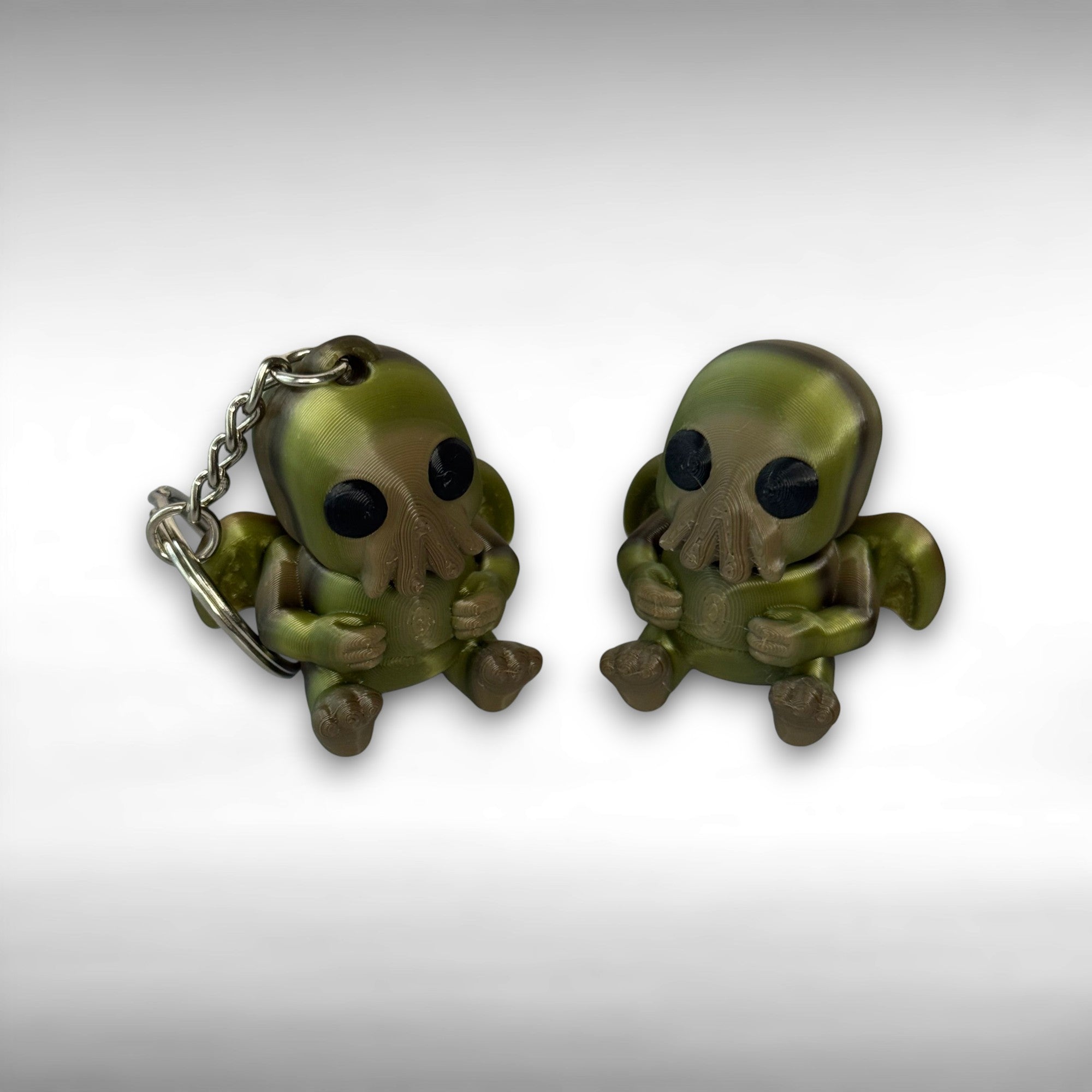Two sitting Cthulhu-shaped figures with wings and face-tentacles, one with keychain and one without, on a gray background