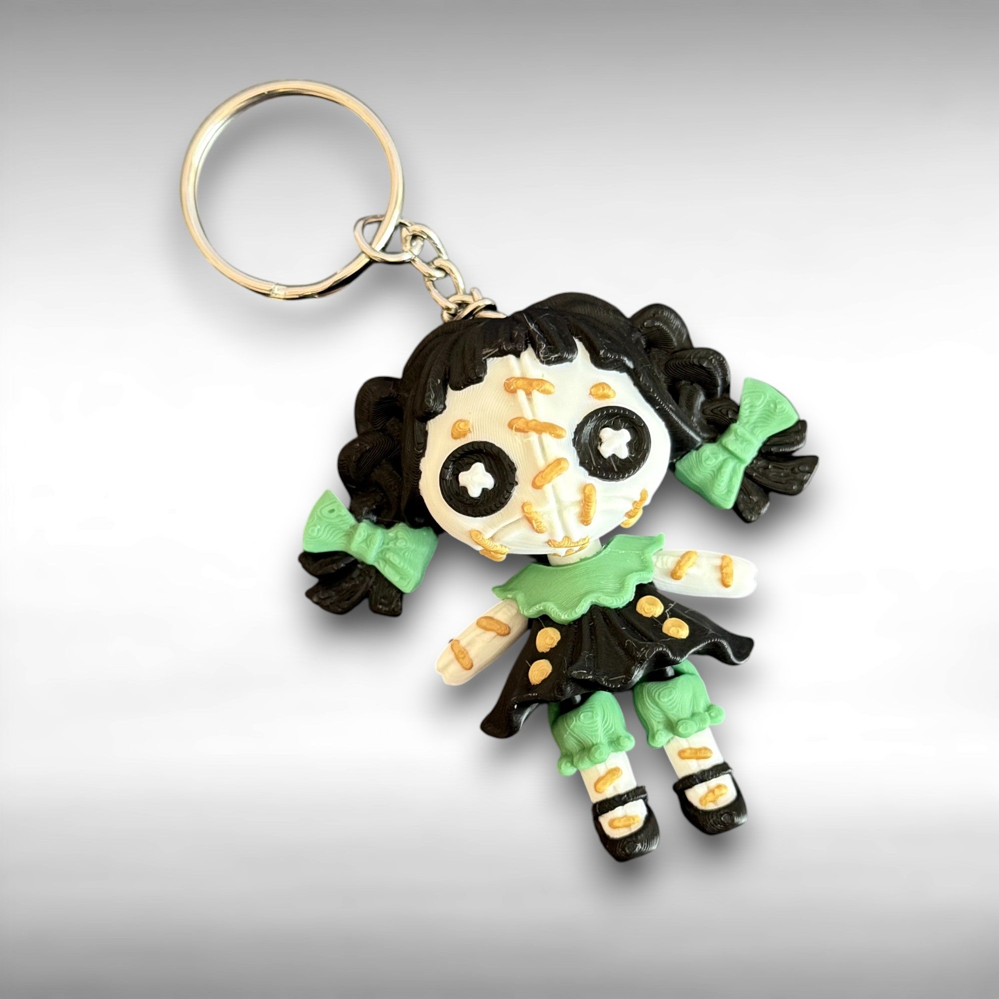 Keychain of a doll character with black hair, button eyes, and a black and green dress on a gray background