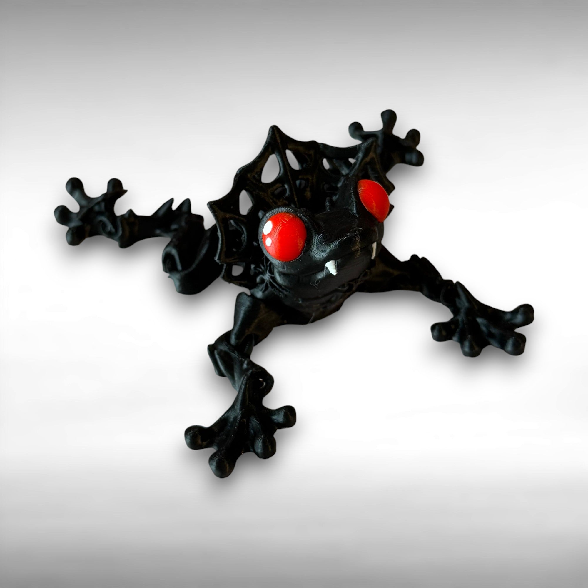 3D printed black and red frog with red eyes and vampire fangs on a gray background