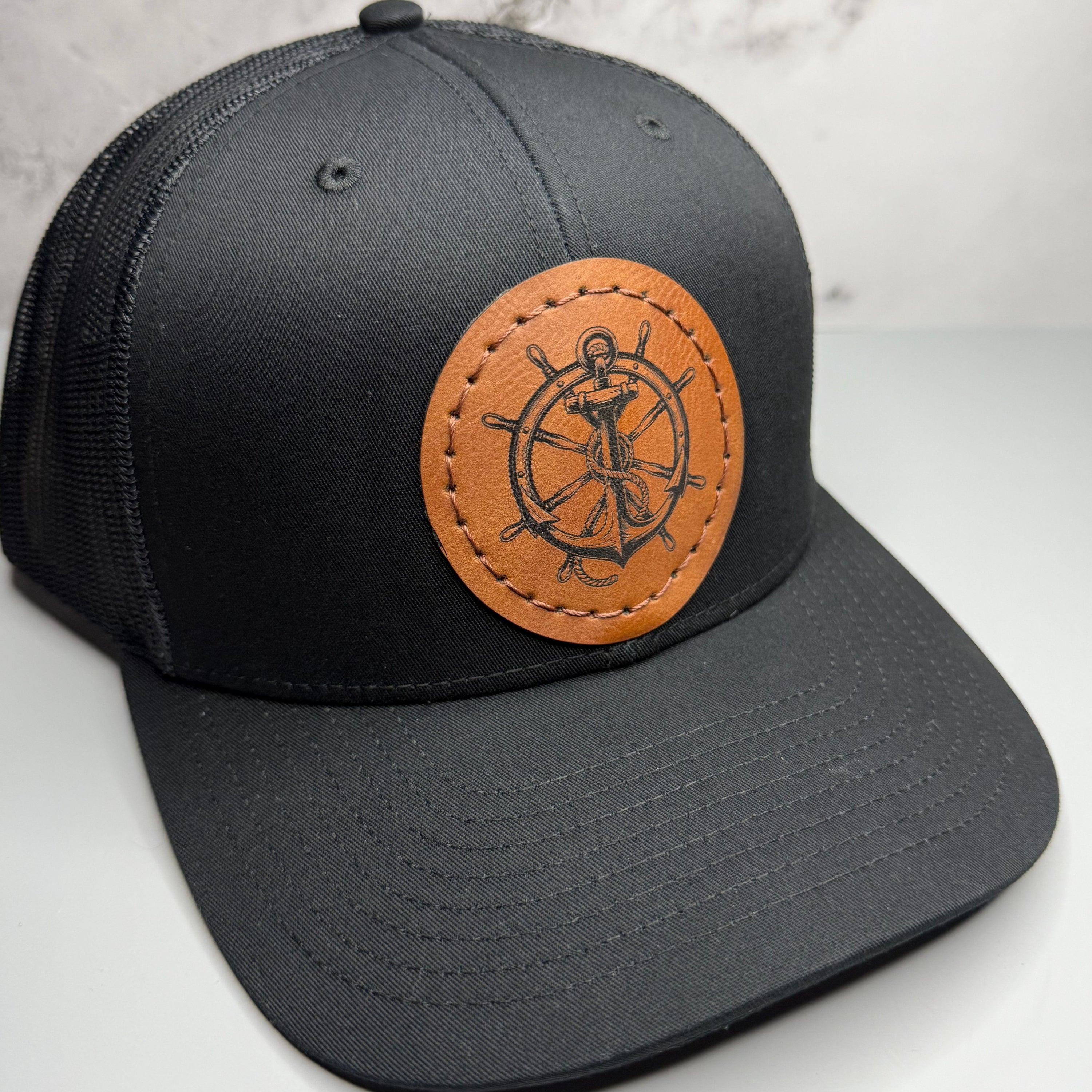 Black cap with a brown leather patch featuring a ship's wheel and anchor 
design on a light gray background.