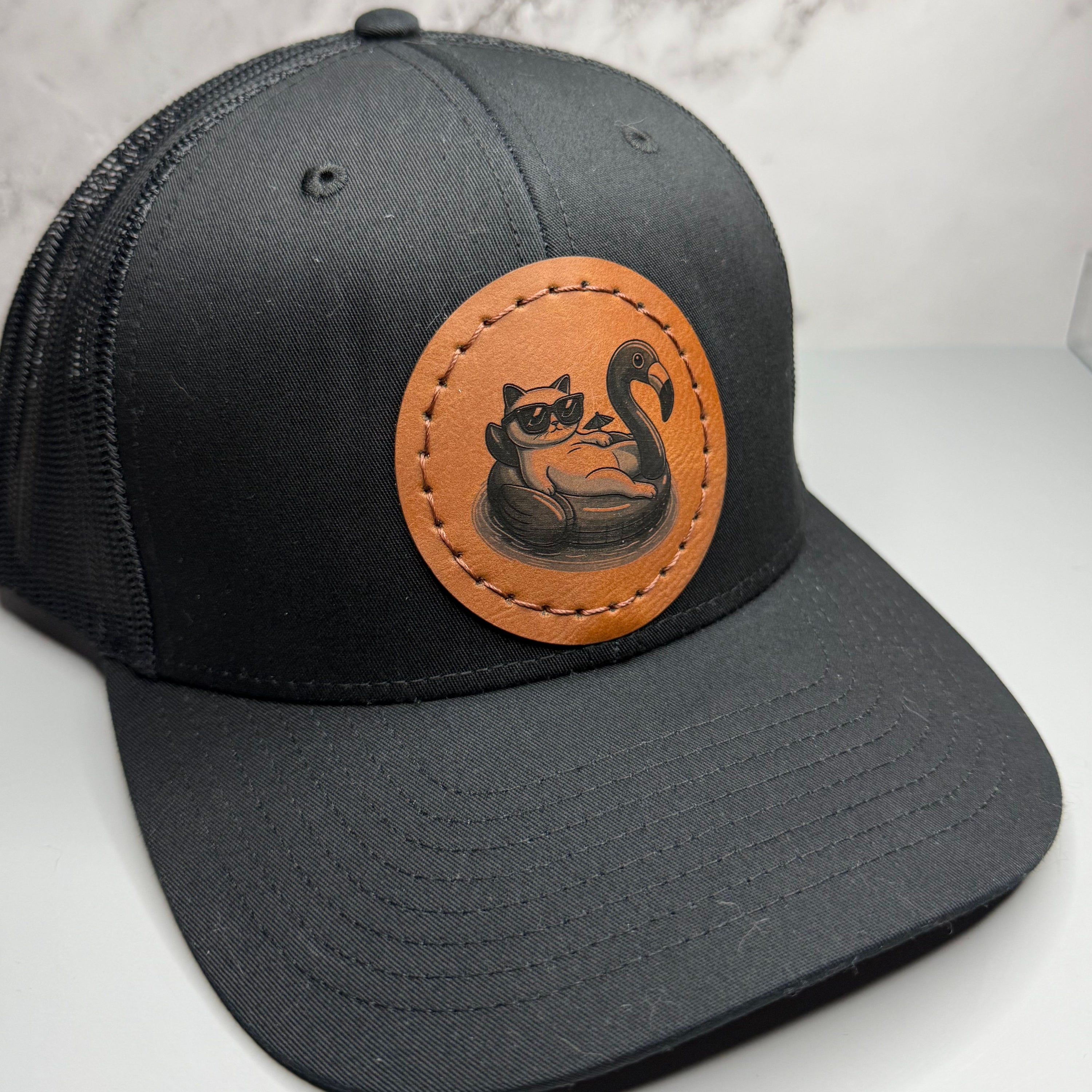 Black cap with a brown leather patch featuring a cat with sunglasses on a flamingo pool float on a gray background