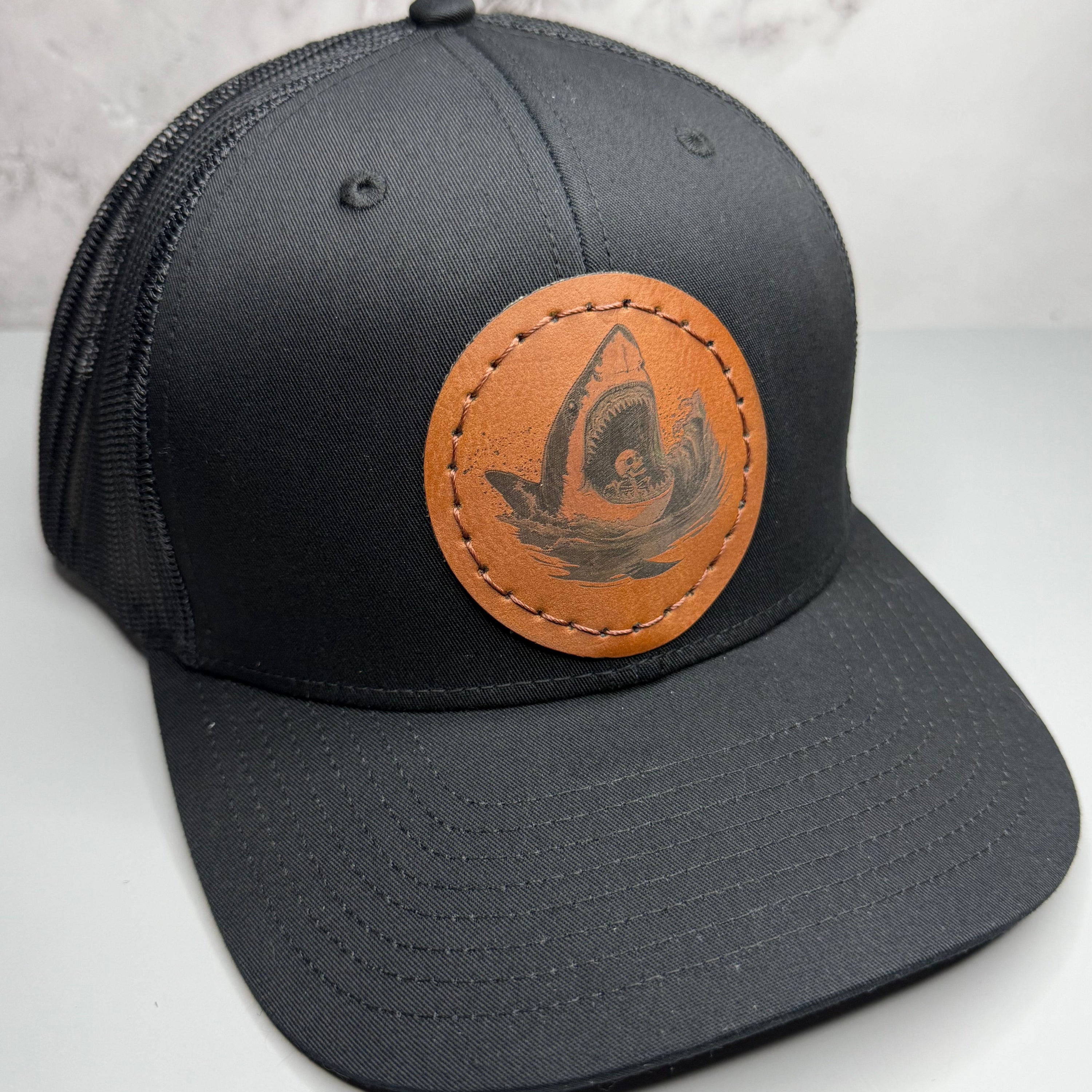 Black trucker hat with a brown leather patch featuring a shark and skeleton design on a gray background